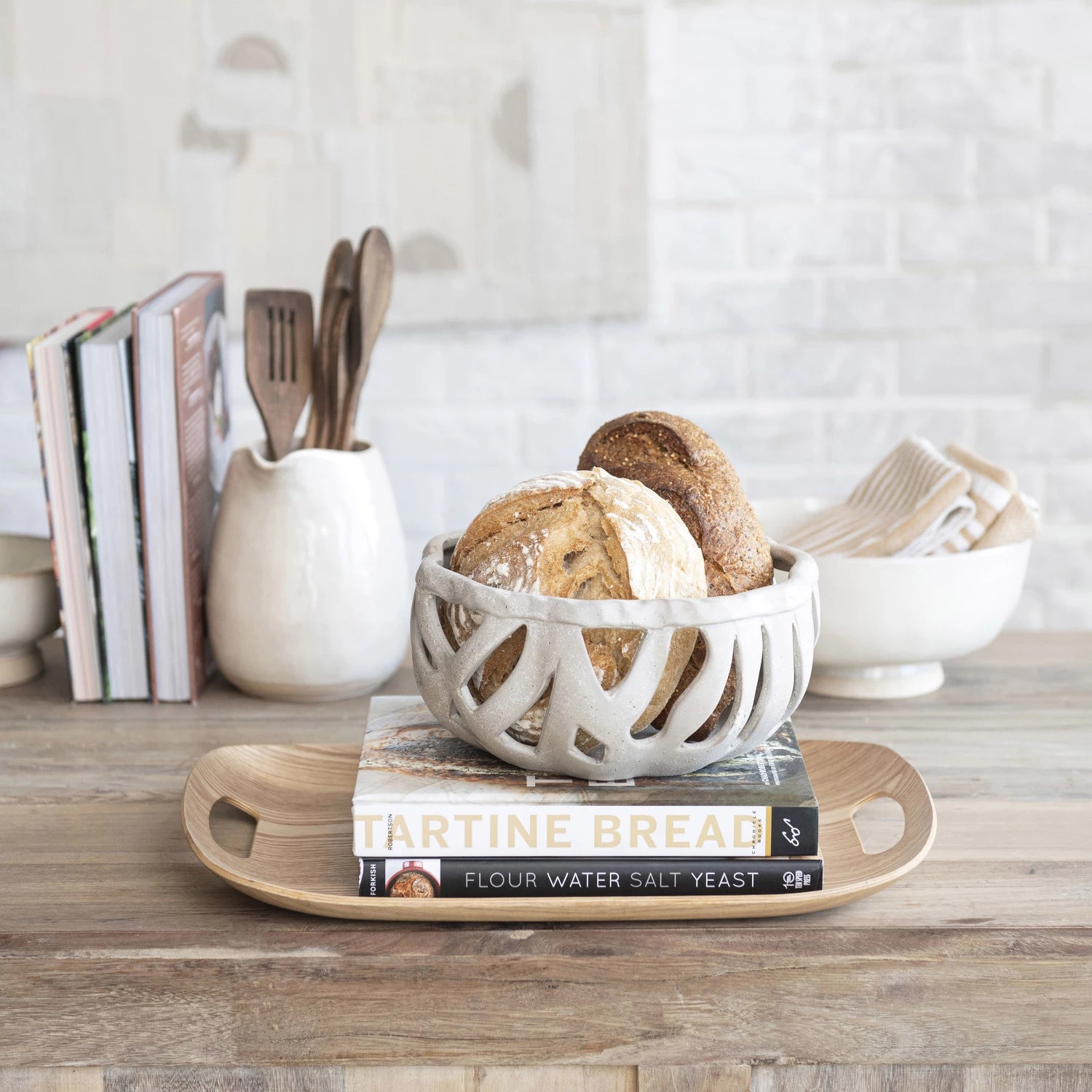 A rustic bungalow-style kitchen setting featuring a ceramic bowl with fresh bread on a Bloomingville Natural Oak Wood Serving Tray, which rests atop a &quot;Tartine Bread&quot; cookbook. Cooking utensils and books are in the background on a wooden counter.