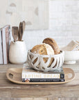 A rustic bungalow-style kitchen setting featuring a ceramic bowl with fresh bread on a Bloomingville Natural Oak Wood Serving Tray, which rests atop a "Tartine Bread" cookbook. Cooking utensils and books are in the background on a wooden counter.