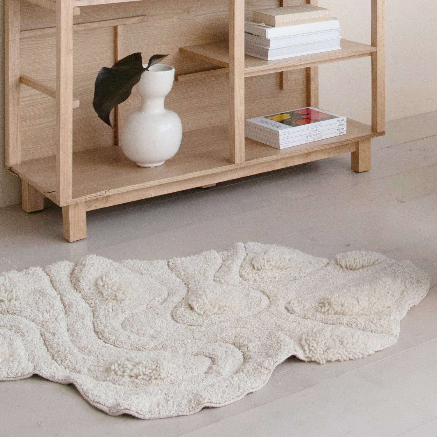 A 3'x3'8" tufted rug by Bloomingville with a wavy texture sits on a light floor in front of a wooden shelf that holds a white vase with a leaf and stacked books, creating a softly lit, minimalist scene.