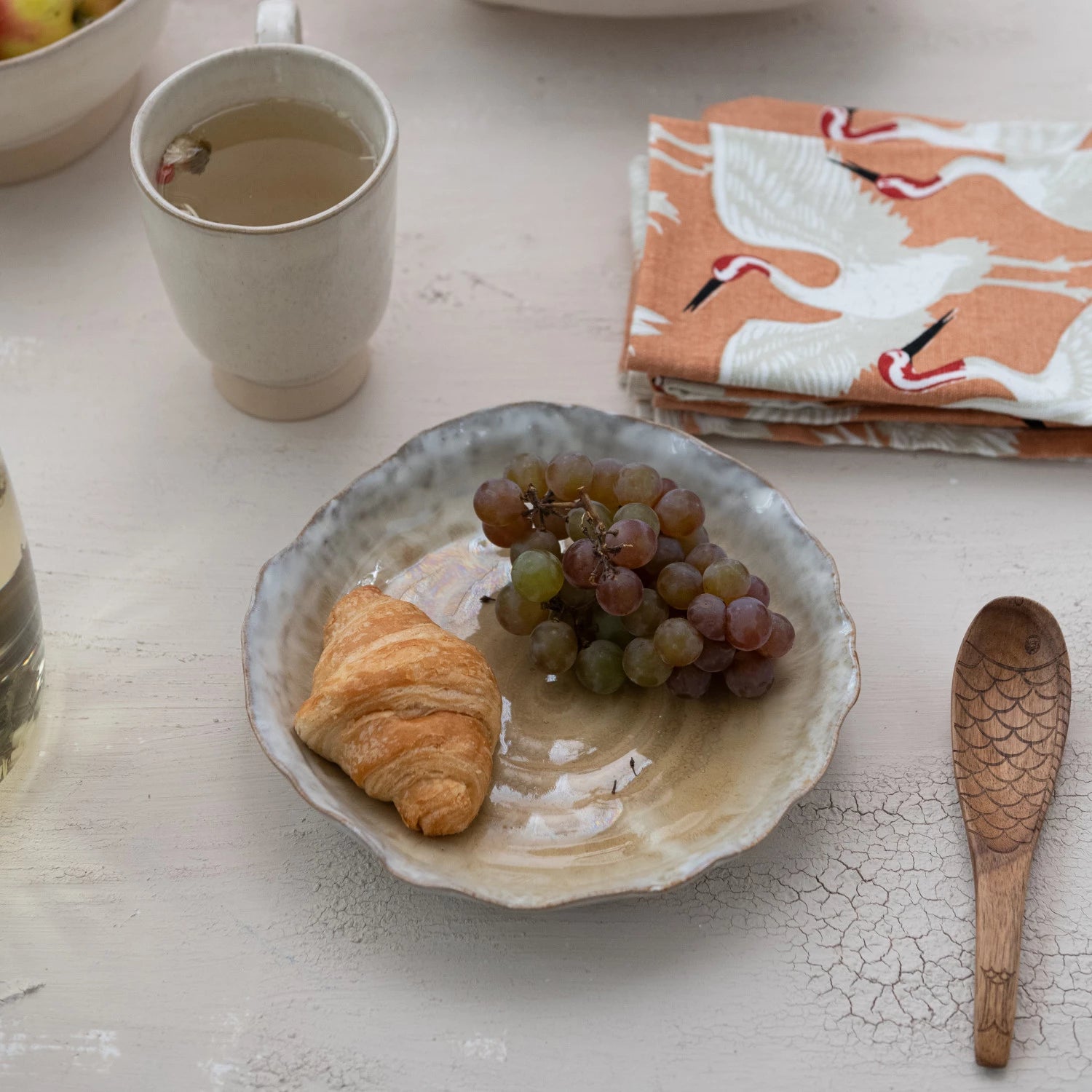 A Creative Co-op Organic Shaped Plate with a croissant and grapes sits on a light table beside tea, a wooden spoon, and a folded orange crane napkin. The plate’s reactive glaze lends subtle elegance to the breakfast scene.