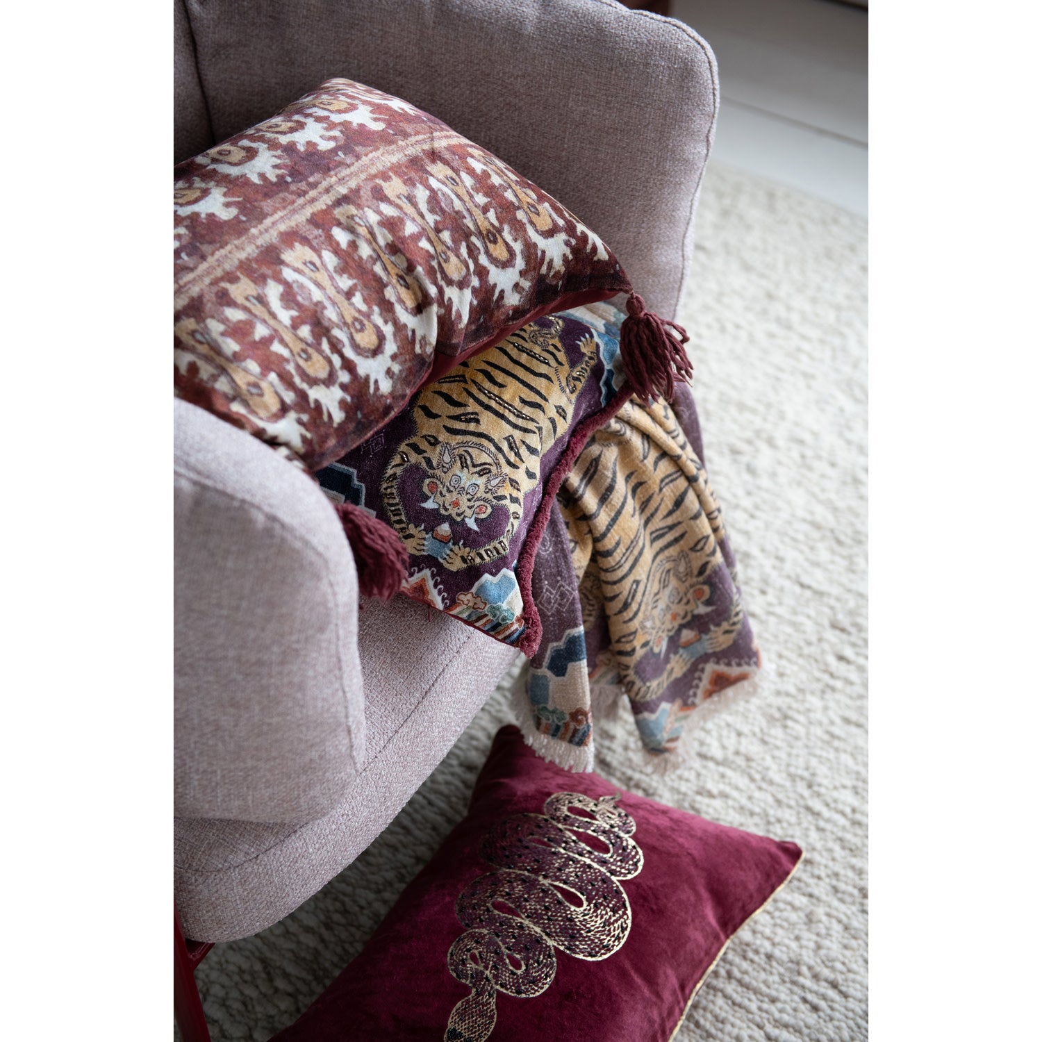 A beige armchair holds a patterned cushion, with a Bloomingville Cotton Throw/Tibetan Tigers draped over the arm. A maroon pillow with gold embroidery lies on the light carpet—a perfect boho decor accent.