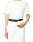 A person dresses in a plain white t-shirt and white pants, accessorized with Jerome Dreyfuss's Gaucho Ceinture belt, crafted from black cowskin calfskin and adorned with domed metal rivets. The minimalist background enhances the ensemble.