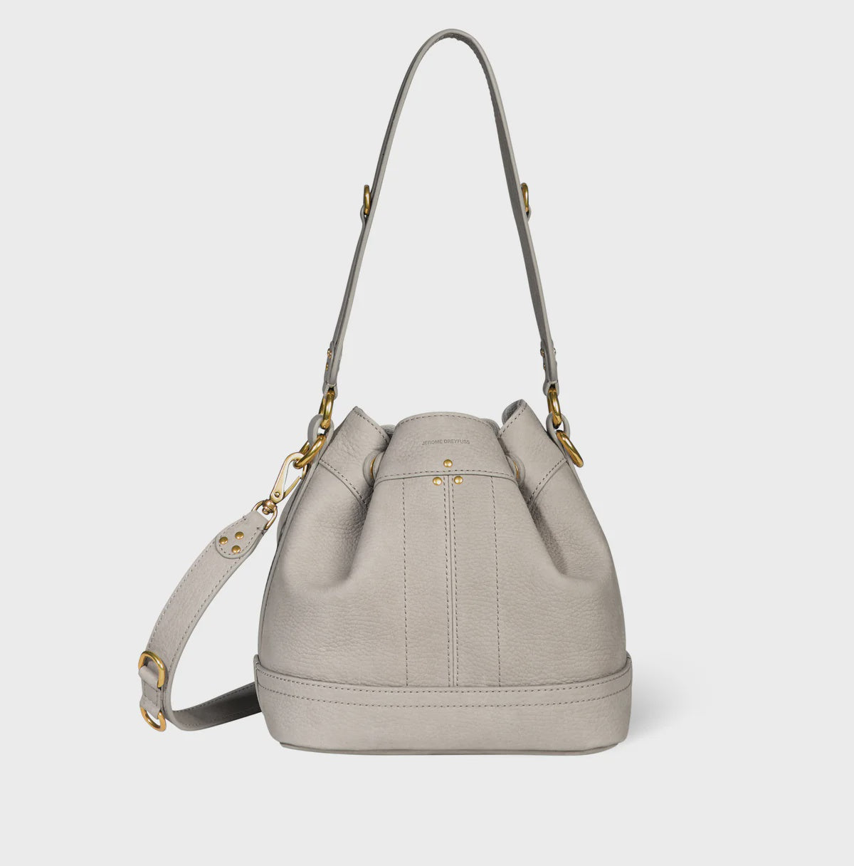 The Ben Small Bucket by Jerome Dreyfuss is a light gray calfskin bag with gold-tone hardware and an adjustable strap, featuring seam details, a drawstring closure, and a minimalist design on a white background.