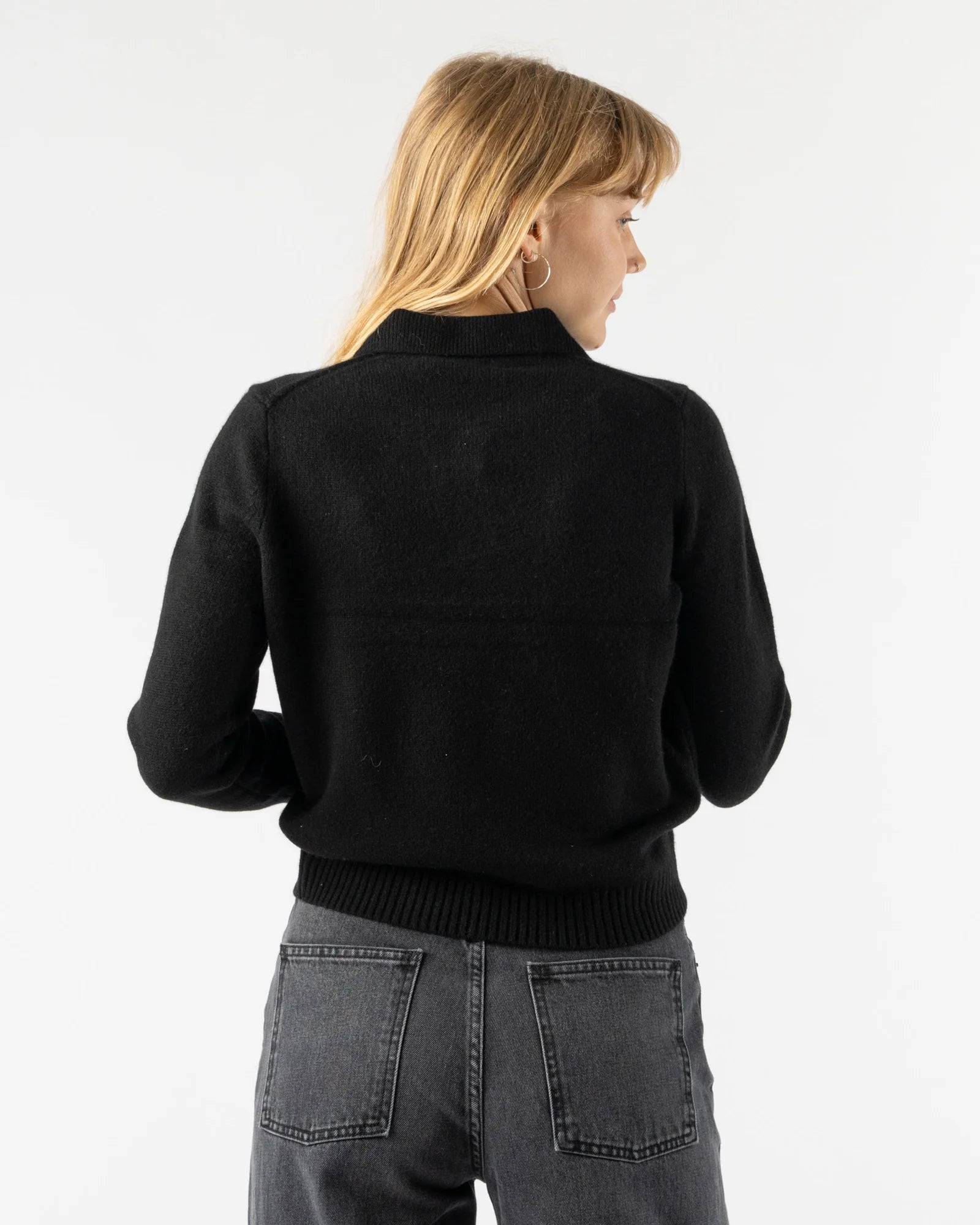 A woman with straight blonde hair, wearing the Alex Mill Alice Polo Cashmere sweater in black and gray jeans, stands facing away from the camera against a plain white background.