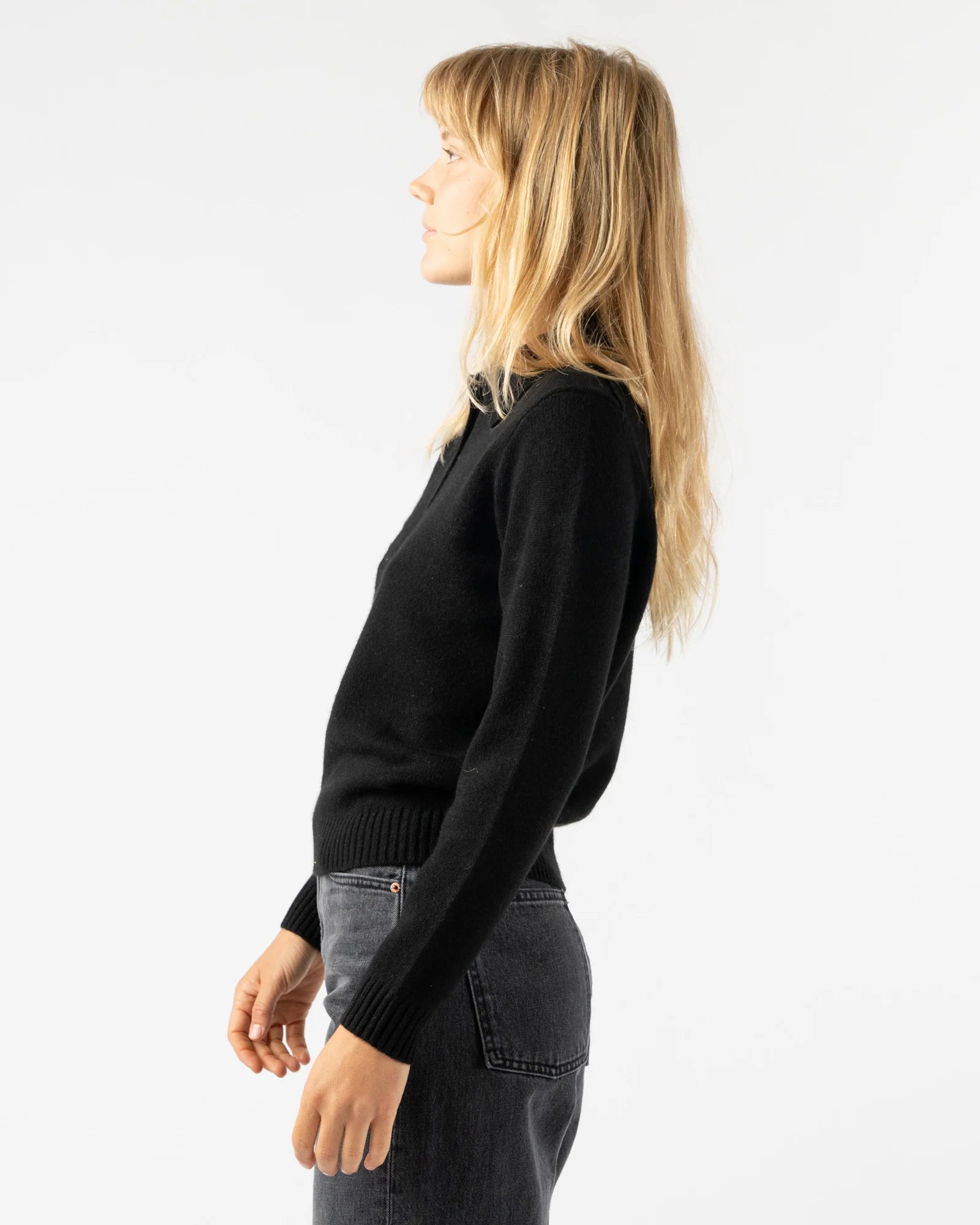 A woman with long blonde hair, wearing the Alex Mill Alice Polo Cashmere sweater and dark jeans, stands in profile against a plain white background.