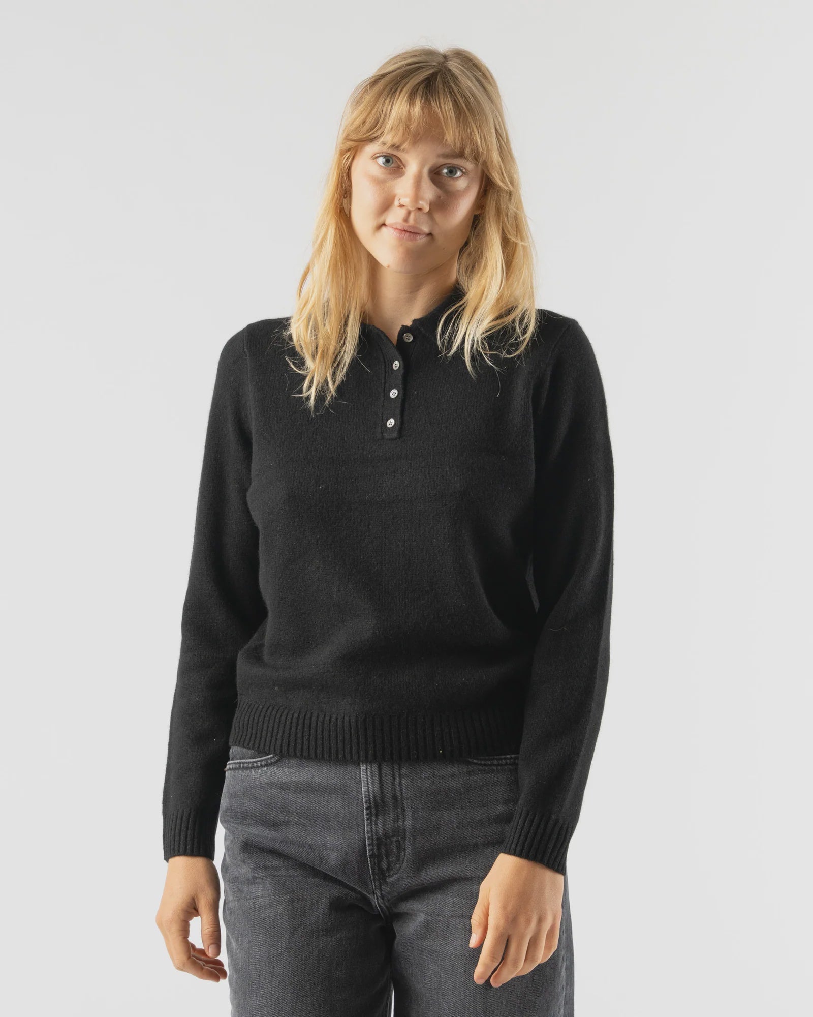 A woman with straight blonde hair wears the Alex Mill Alice Polo Cashmere sweater in black and dark gray jeans, standing against a plain light background with a neutral expression.