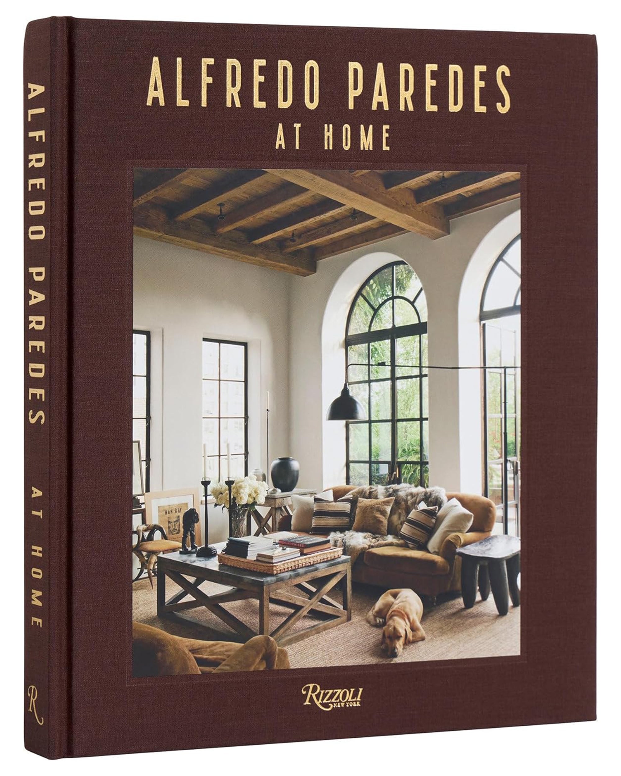The cover of "At Home; Alfredo Paredes" by Random House shows a chic living room with arched windows, wooden beams, elegant furnishings, and a dog on the floor—capturing the designer's modern glamour.