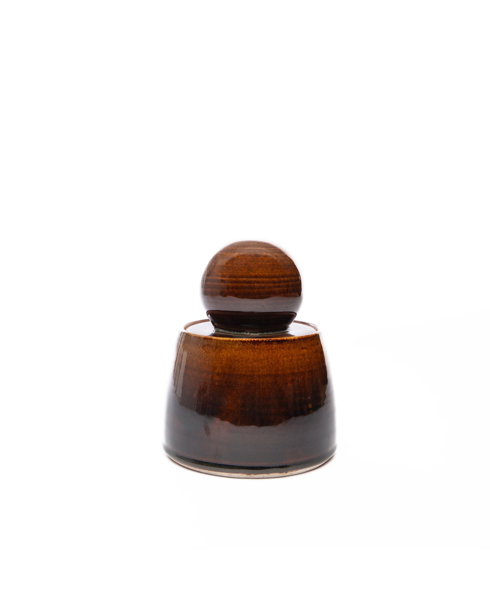 The Faire Amber Stash Jar is a small, brown ceramic container with a glossy amber glaze and a matching rounded lid, shown on a plain white background.