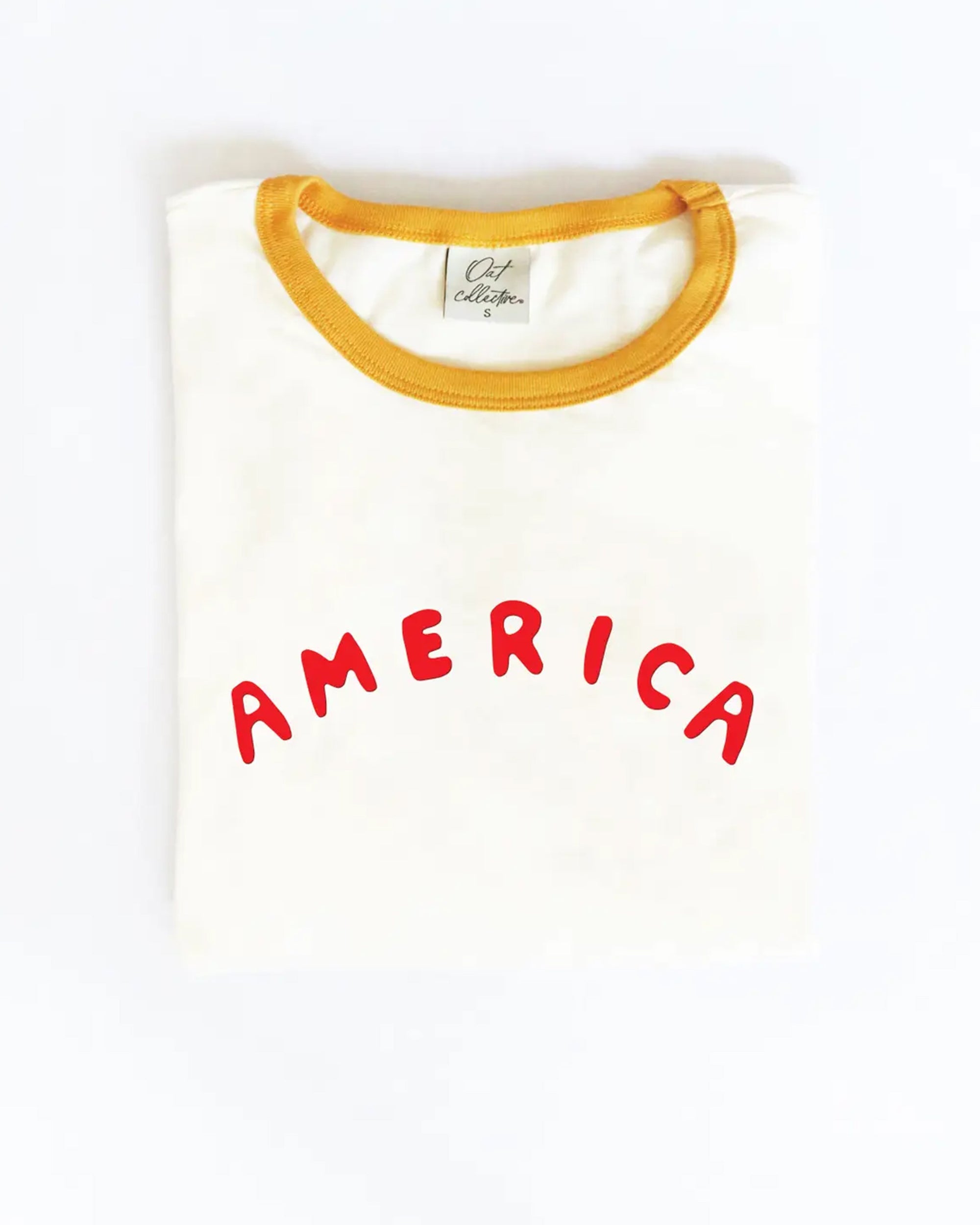 The America Puff Ringer Graphic T-Shirt by Oat Collective is a folded white tee with yellow trim, made from premium Airlume cotton and featuring "AMERICA" in bold red puff print on the front.