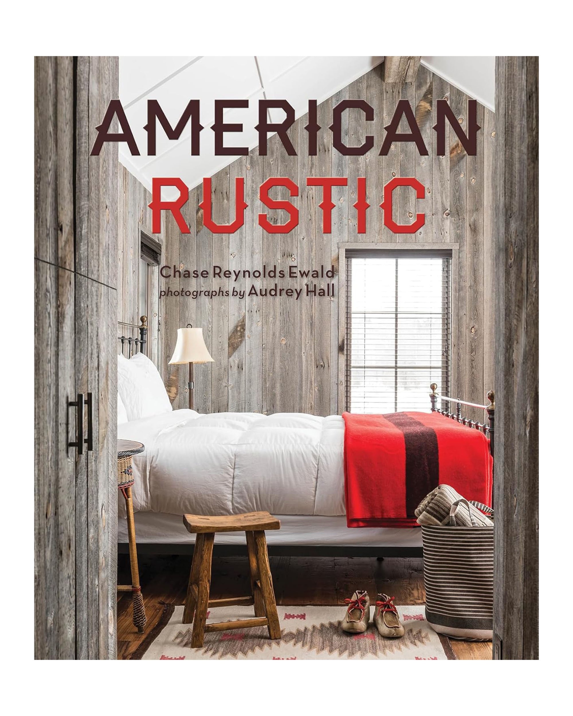 Cover of "American Rustic" by Gibbs Smith Publisher, showing a cozy American rustic bedroom with wood-paneled walls, a white bed with red and black blanket, wooden stools, woven basket, and sunlight through the window.