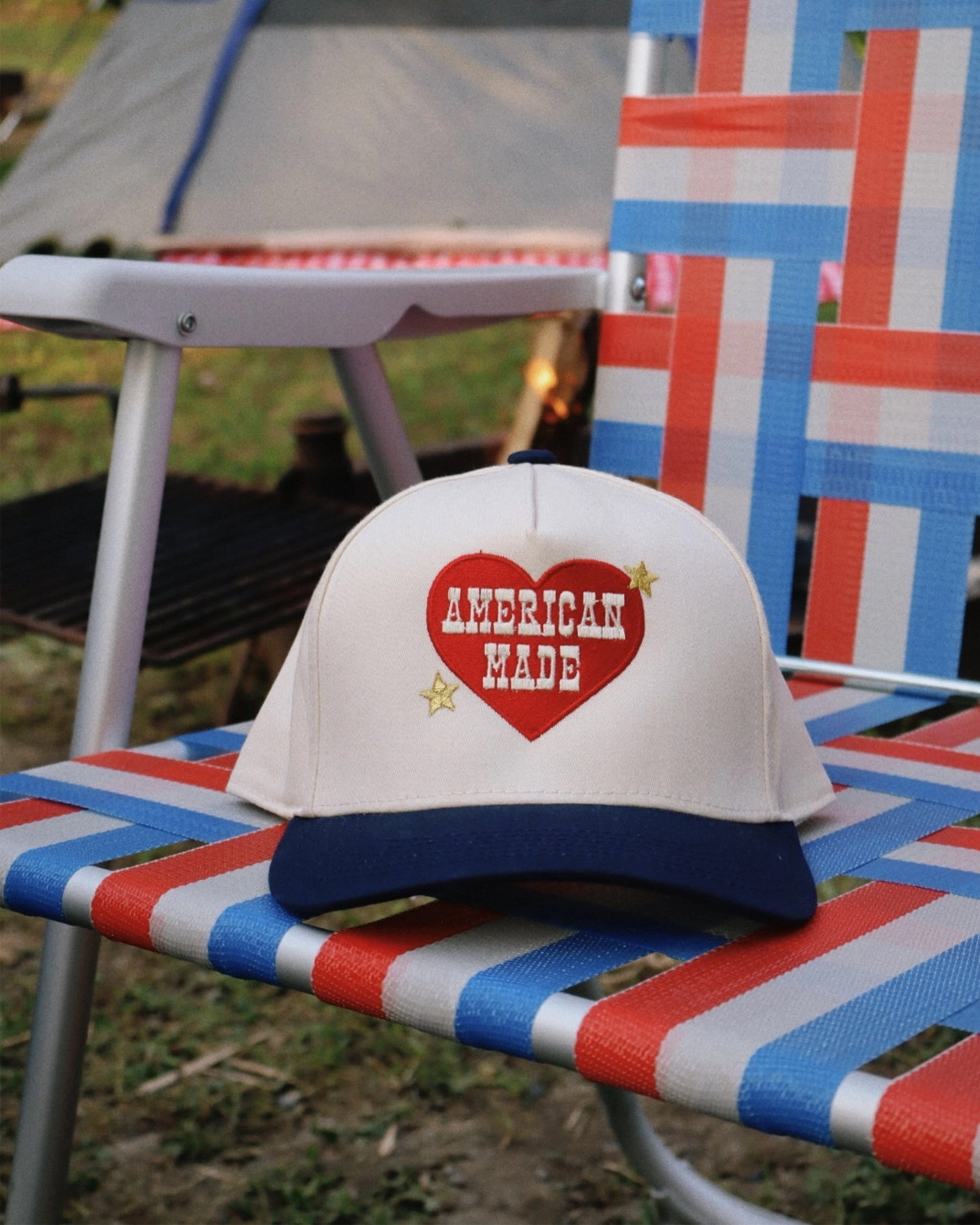 The Faire American Made Trucker Hat, white and blue with a red heart, rests on a red, white, and blue woven lawn chair outdoors amid grass and camping gear. This adjustable snapback is perfect for outdoor adventures.
