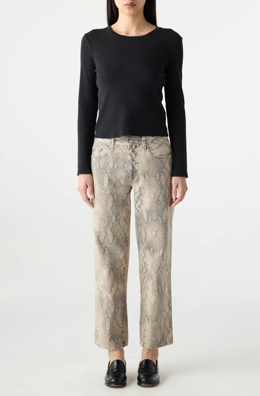 A woman faces forward against a plain white background, wearing a fitted black long-sleeve top, AMO Denim Billie Pants—premium rigid denim cropped wide leg jeans in beige snake-print—and black loafers.