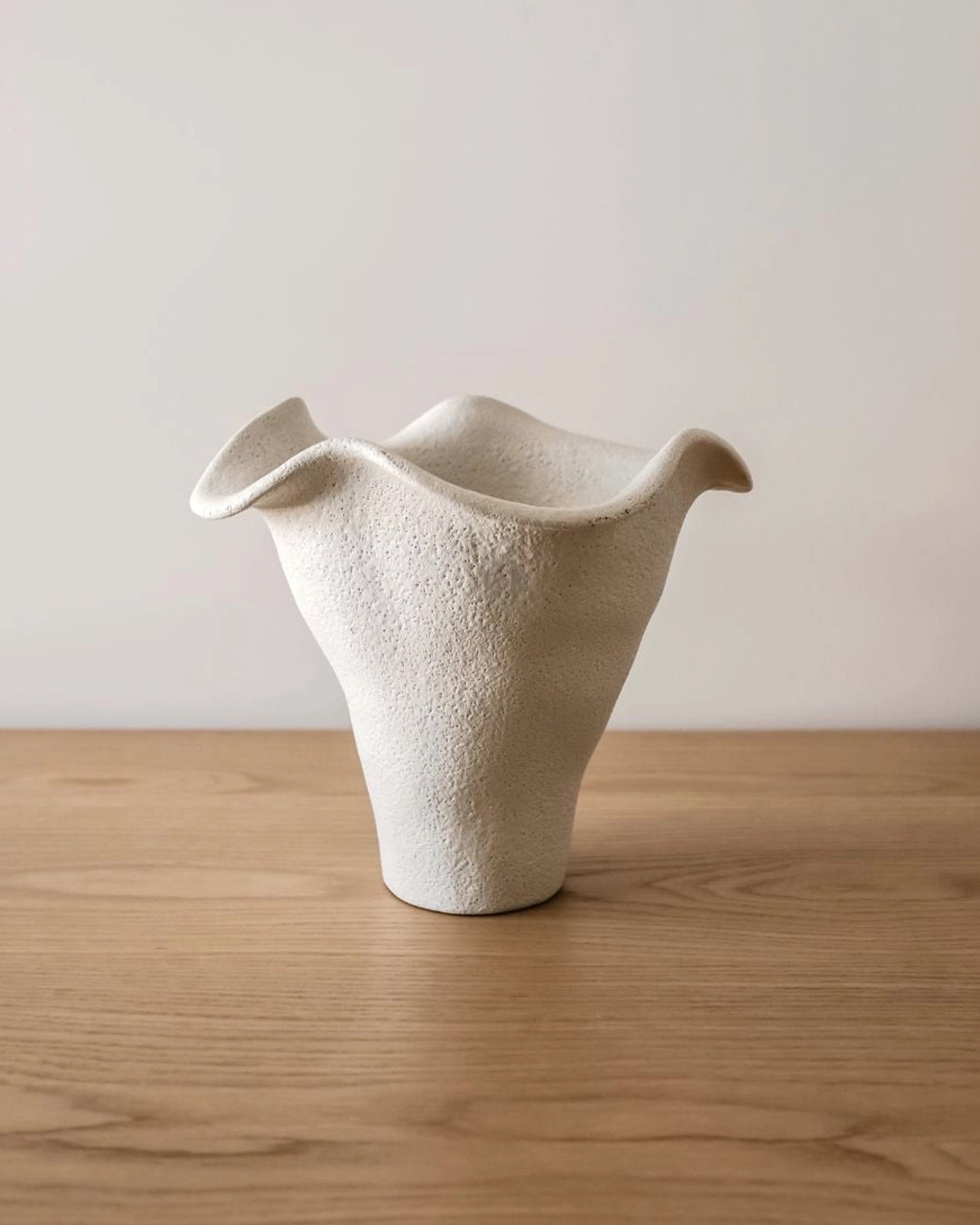 The Faire Andrea Bouquet Vase—a white, textured ceramic piece with an organic, modern shape—rests on a light wood surface against a plain, light background.