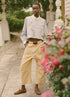 A person stands outdoors on a stone path in Sea New York’s Andrea WorkWear Pants—yellow barrel-leg style—paired with a light blue patterned shirt, black belt, and brown boat shoes. Pink flowers and greenery frame this contemporary scene.