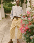 A person stands outdoors on a stone path in Sea New York’s Andrea WorkWear Pants—yellow barrel-leg style—paired with a light blue patterned shirt, black belt, and brown boat shoes. Pink flowers and greenery frame this contemporary scene.