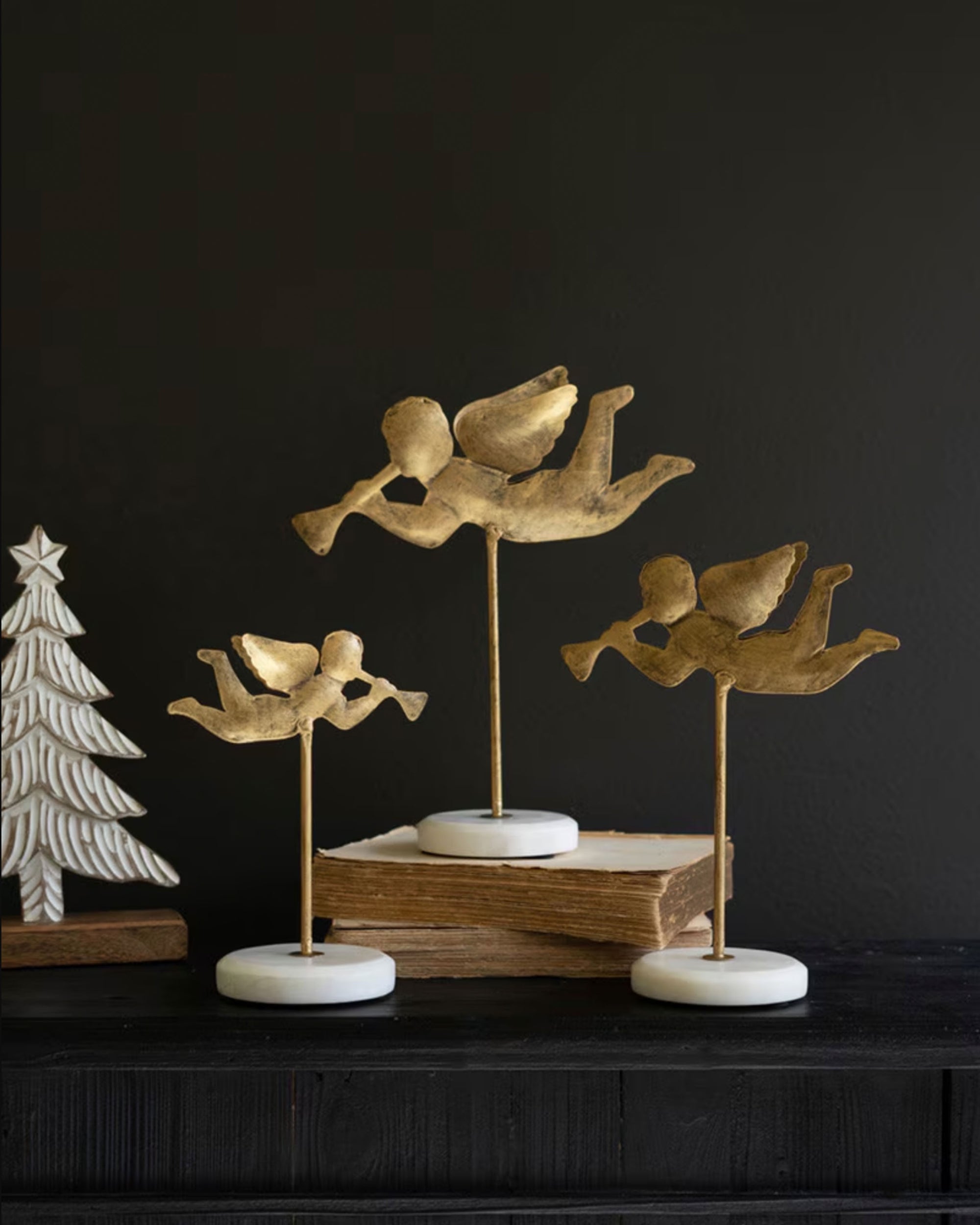 Three Kalalou, Inc Angel On Marble Base figurines—gold angels in flight, each playing a trumpet—are displayed on a dark surface with books, beside a white Christmas tree, creating a striking holiday gift scene.