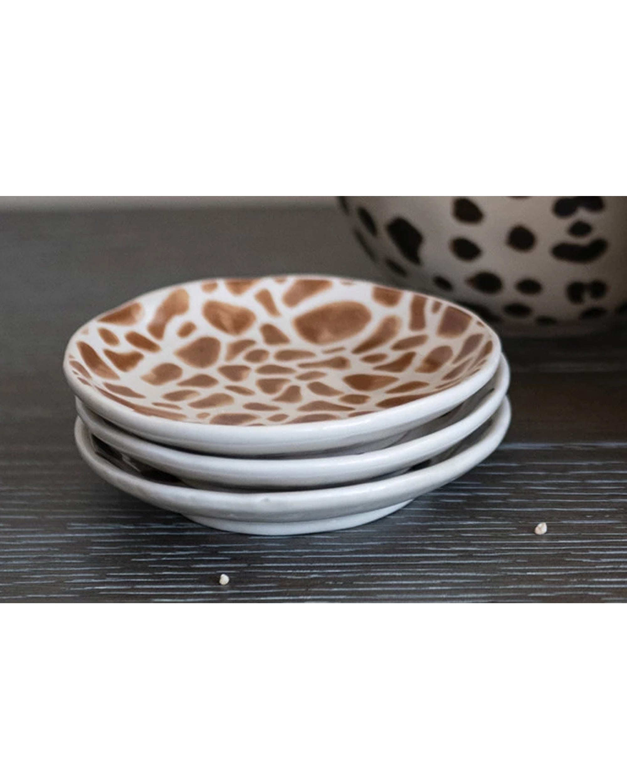 Three small Creative Co-op Dish with Animal Print plates featuring brown giraffe patterns are stacked on a dark wooden surface, with black-and-white spotted dishes partially visible in the background.