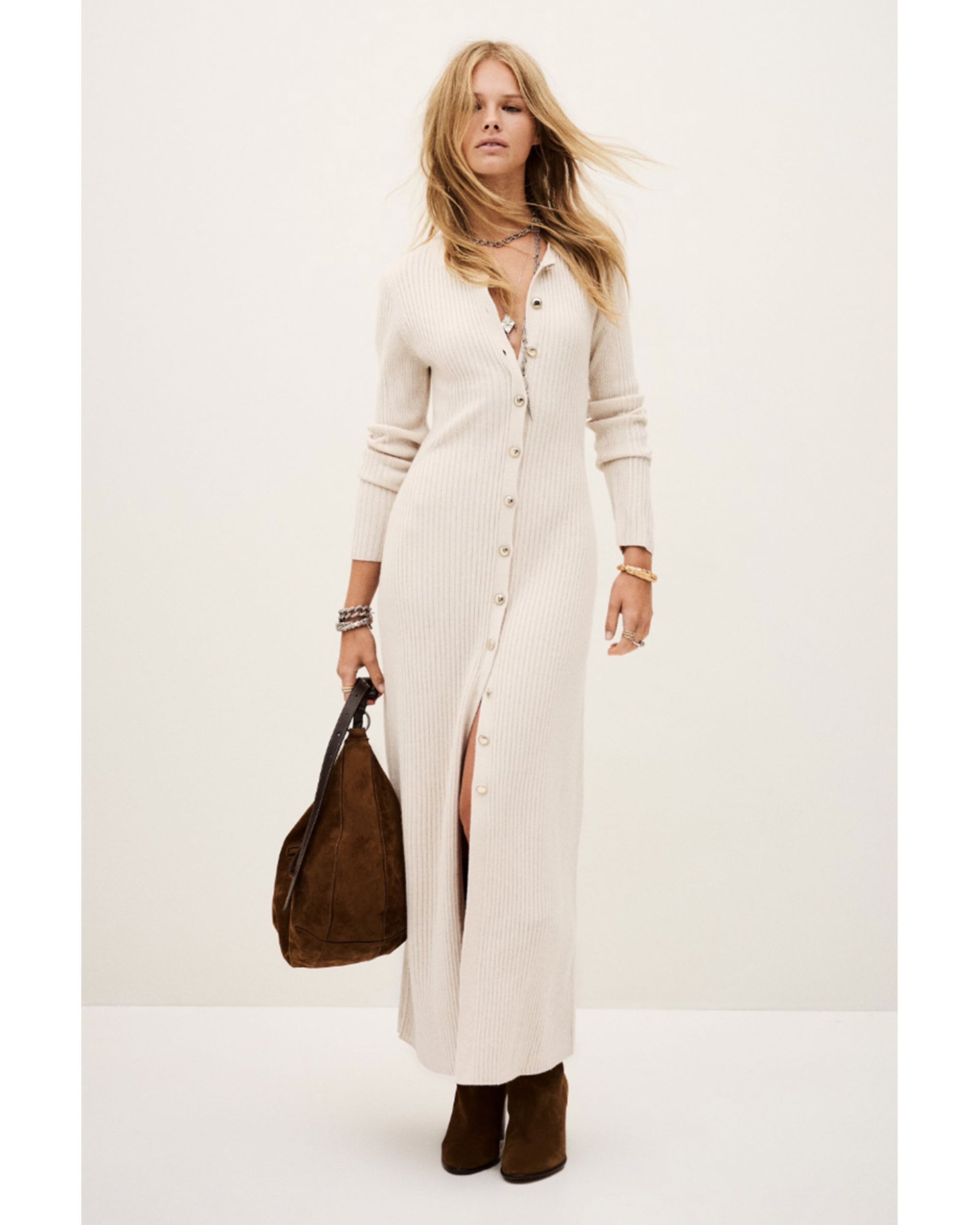 A woman stands against a plain background wearing the BA&SH Dress Anisa—a long, cream ribbed wool blend button-down dress with long sleeves. She pairs it with brown ankle boots, a large brown bag, and has loose wavy blonde hair and minimal jewelry.