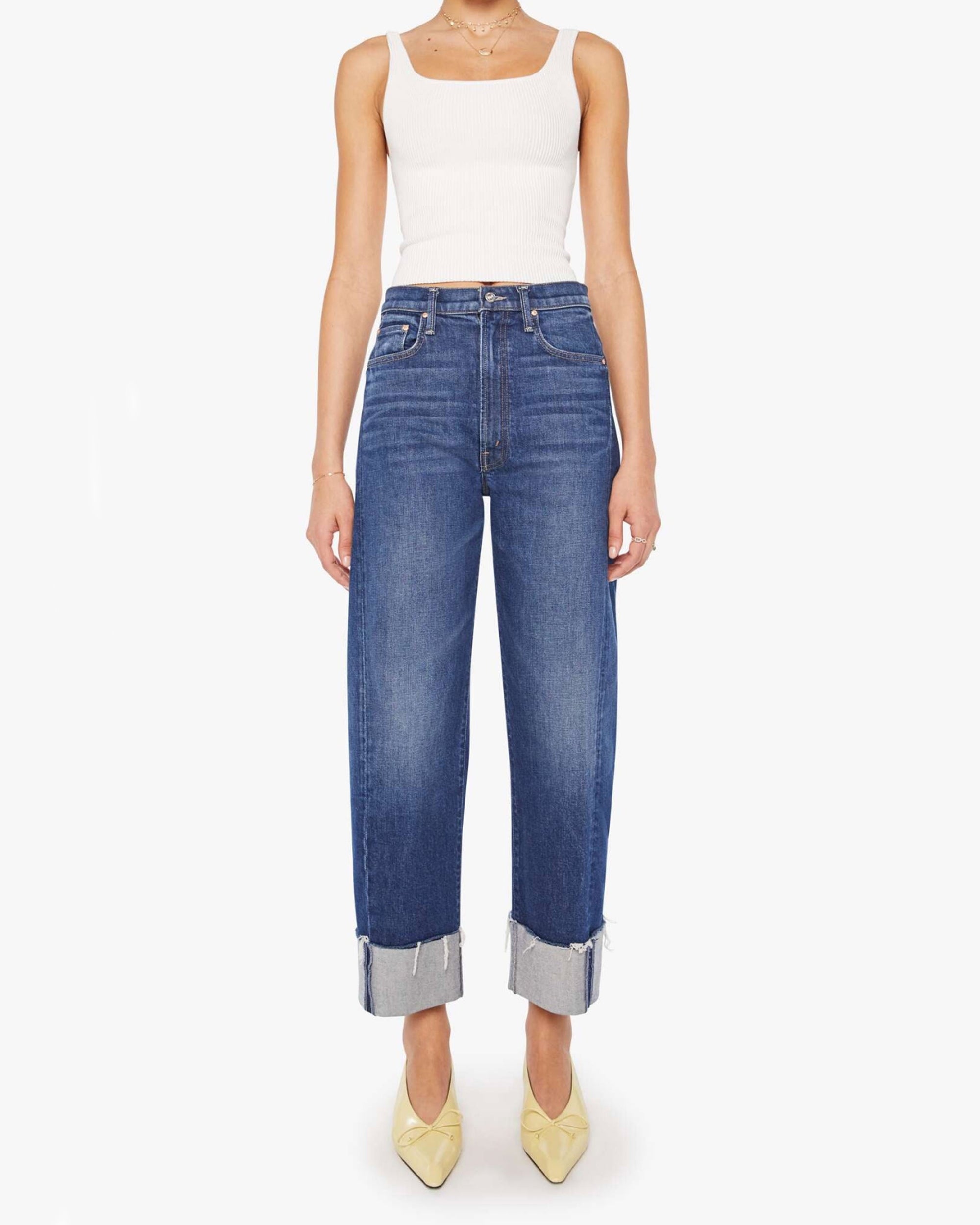 A person stands against a plain white background wearing a white sleeveless ribbed top, Mother’s The Half-Pipe Ankle Cuff Fray high-waisted dark blue barrel jeans with wide cuffs, and pale yellow pointed-toe shoes.