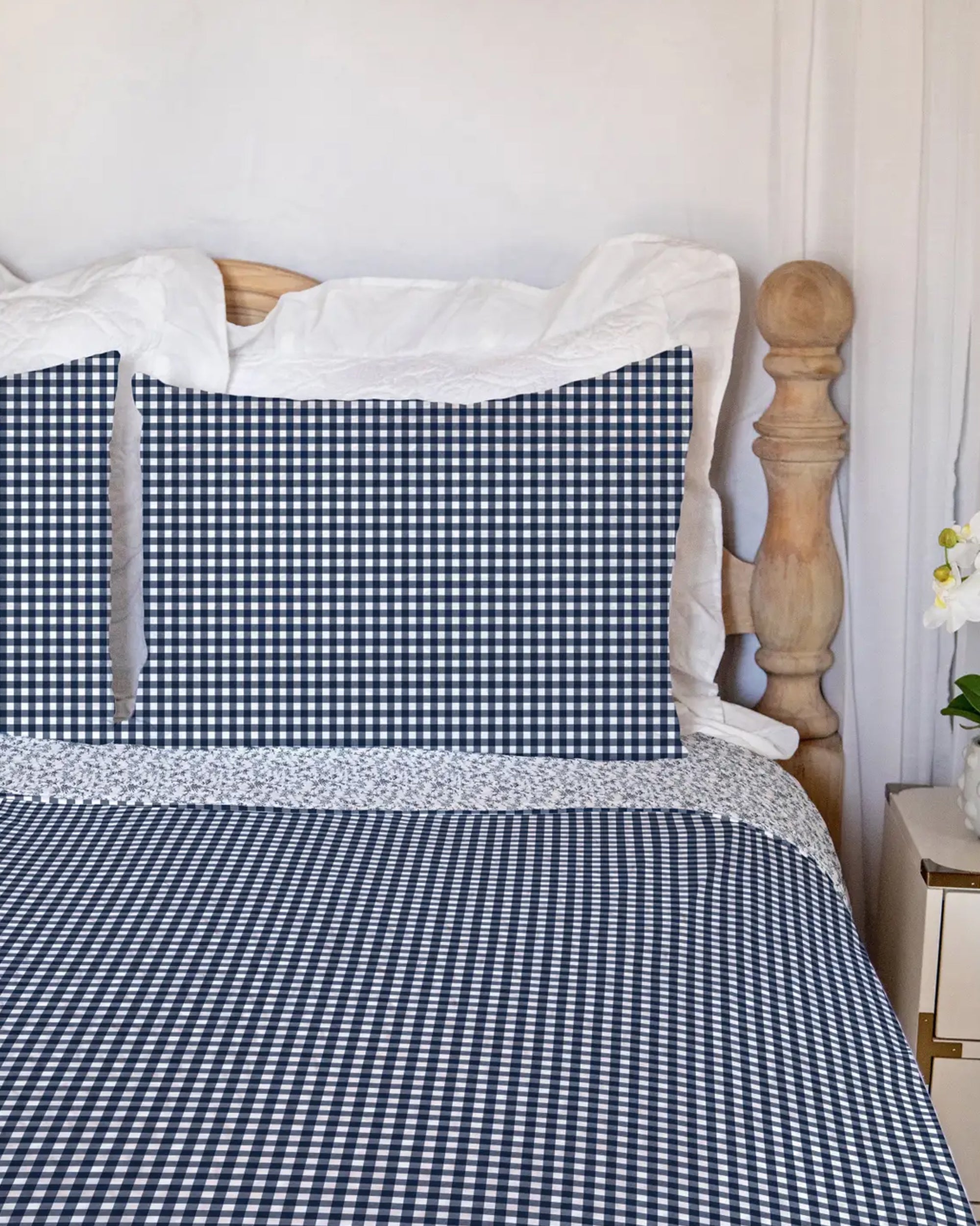 A neatly made bed features the Faire Anthia Gingham Navy Quilt with matching pillows, crisp white sheets, a wooden headboard, and a nightstand topped with white flowers.