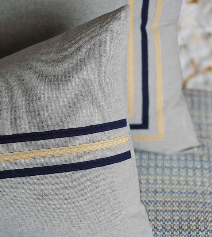 Close-up of two Eastern Accents Sprouse Linear Pillows in pewter with ribbon trim—one featuring parallel navy and beige stripes, the other bordered in navy and beige—resting on a textured, patterned surface.