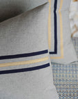 Close-up of two Eastern Accents Sprouse Linear Pillows in pewter with ribbon trim—one featuring parallel navy and beige stripes, the other bordered in navy and beige—resting on a textured, patterned surface.