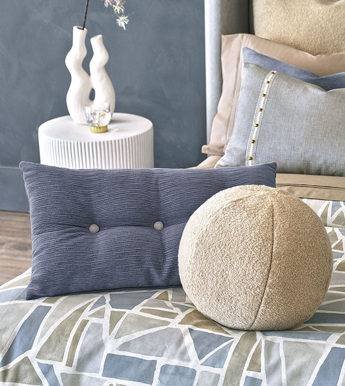 A modern bedroom features the APG-498 OLIVER BOUCLE by Eastern Accents—a spherical boucle pillow—on a geometric bedspread, paired with a blue rectangular cushion and a white side table with a sculptural vase and candle.