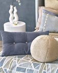 A modern bedroom features the APG-498 OLIVER BOUCLE by Eastern Accents—a spherical boucle pillow—on a geometric bedspread, paired with a blue rectangular cushion and a white side table with a sculptural vase and candle.
