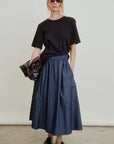 Wearing sunglasses, a loose black t-shirt, Aquarius Cocktail’s Le Cargo Skirt in blue Japanese cotton poplin with oversized pockets, and black slip-on shoes, this person stands against a plain background holding a shiny black bag.
