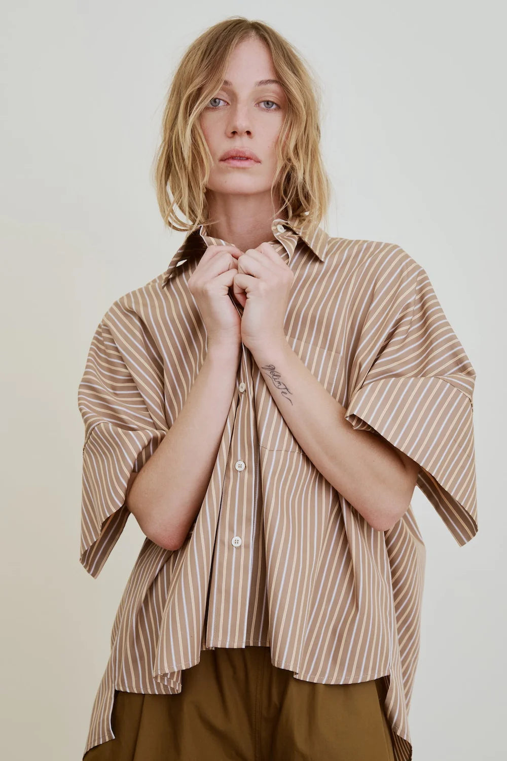 A person with shoulder-length blonde hair wears the Aquarius Cocktail Vesper OS Shirt, a cropped boxy blouse with brown and white stripes and wide sleeves, paired with brown pants against a plain light background.