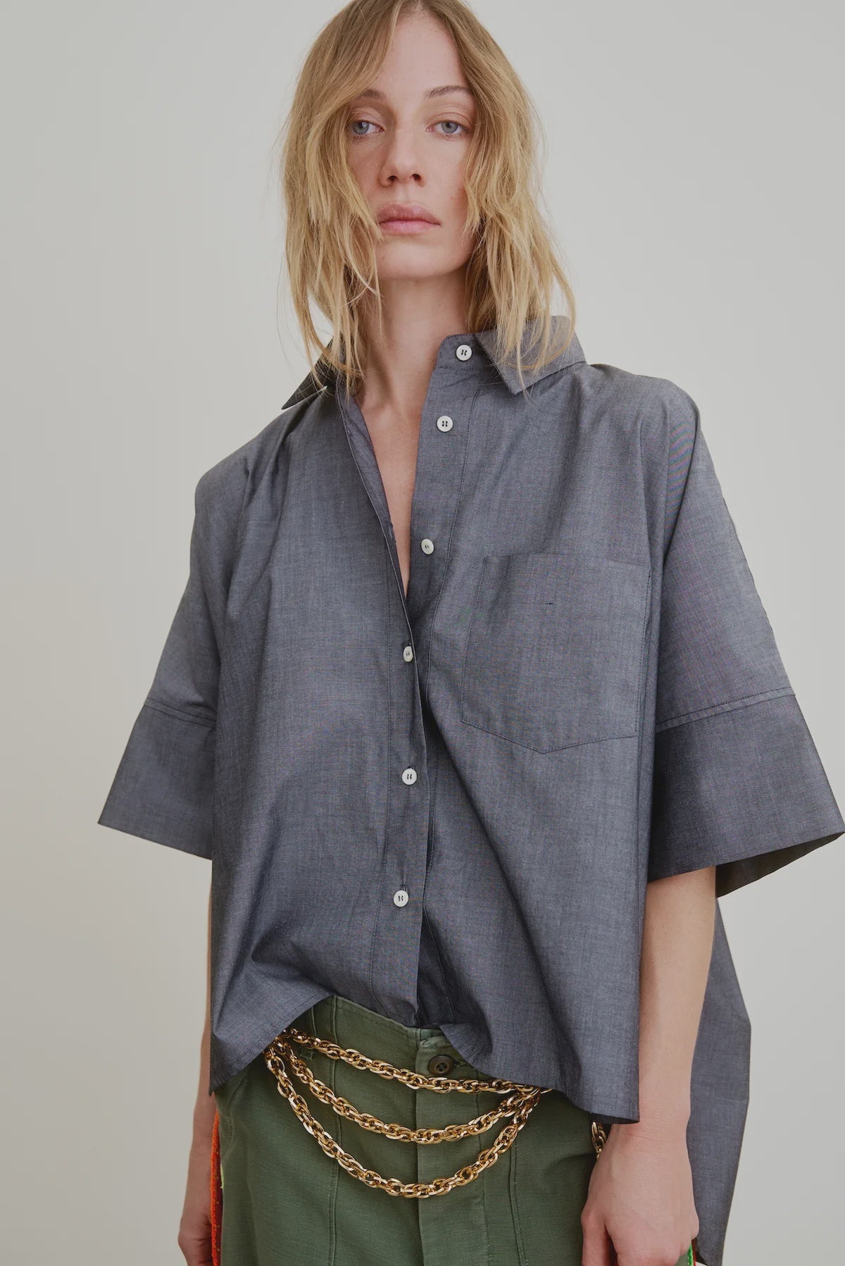 A person with tousled blonde hair wears the Aquarius Cocktail Vesper OS Shirt— a cropped boxy chambray cotton blouse with a front pocket—paired with green pants and layered gold chains, standing against a plain background.