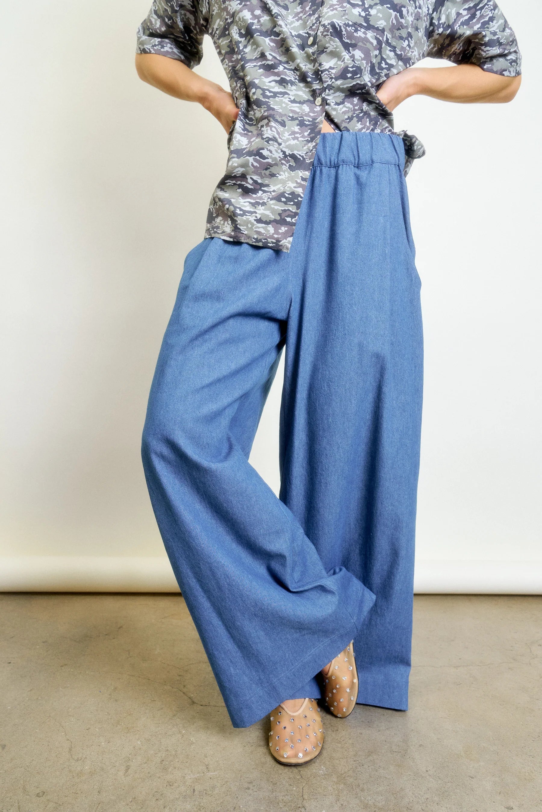 A person wears Aquarius Cocktail’s Connor Wide Leg Pant in blue with a short-sleeved gray camo top and beige studded shoes, standing on a concrete floor. Their face is not visible.