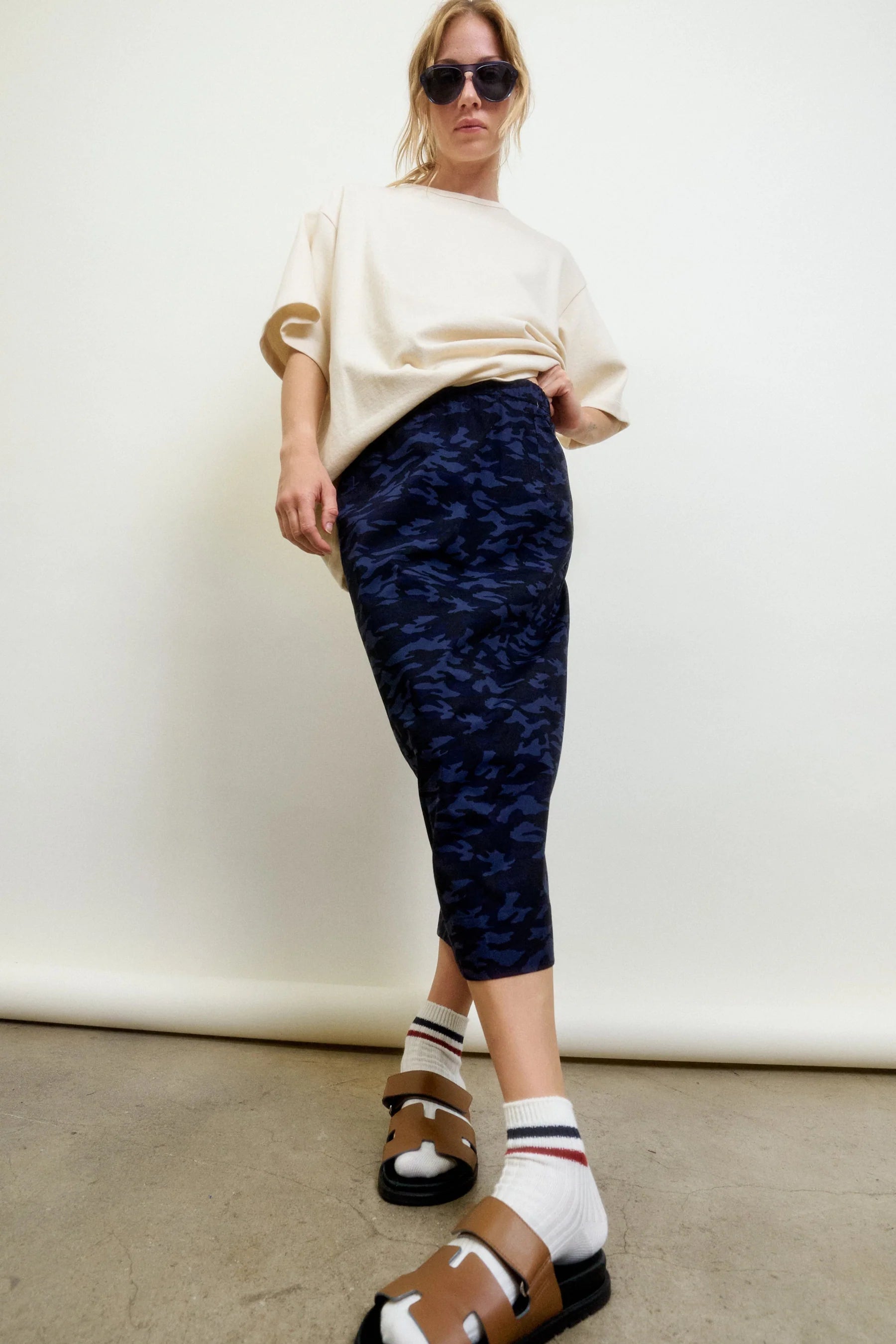 A woman wearing sunglasses, a loose beige t-shirt, the Aquarius Cocktail CAMO LE Pencil Slim Skirt, white socks, and brown sandals stands confidently against a plain background.