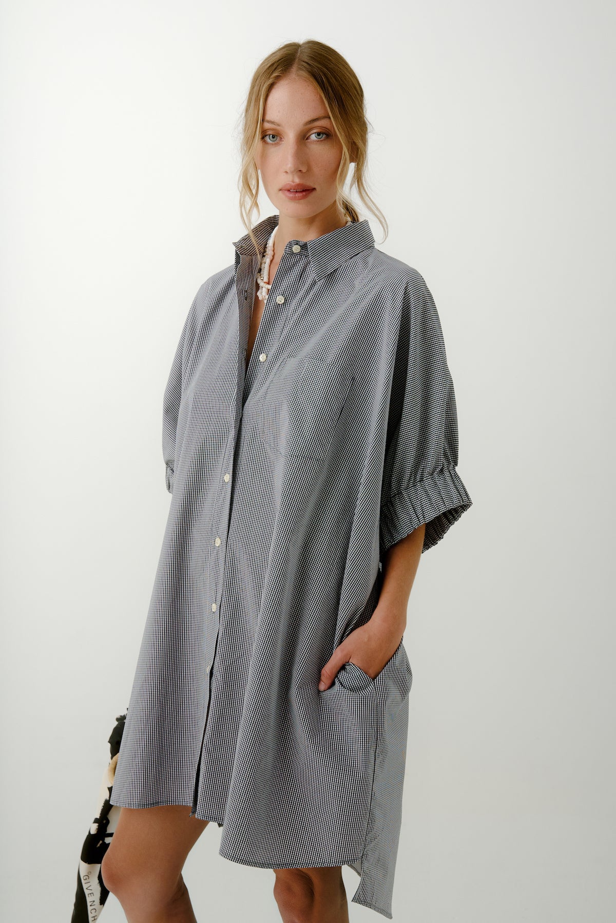 A woman with loose, light brown waves stands against a plain background, wearing the Aquarius Cocktail Le Shorty Oversized Shirt in Japanese poplin with subtle stripes, her hands casually tucked into the pockets.