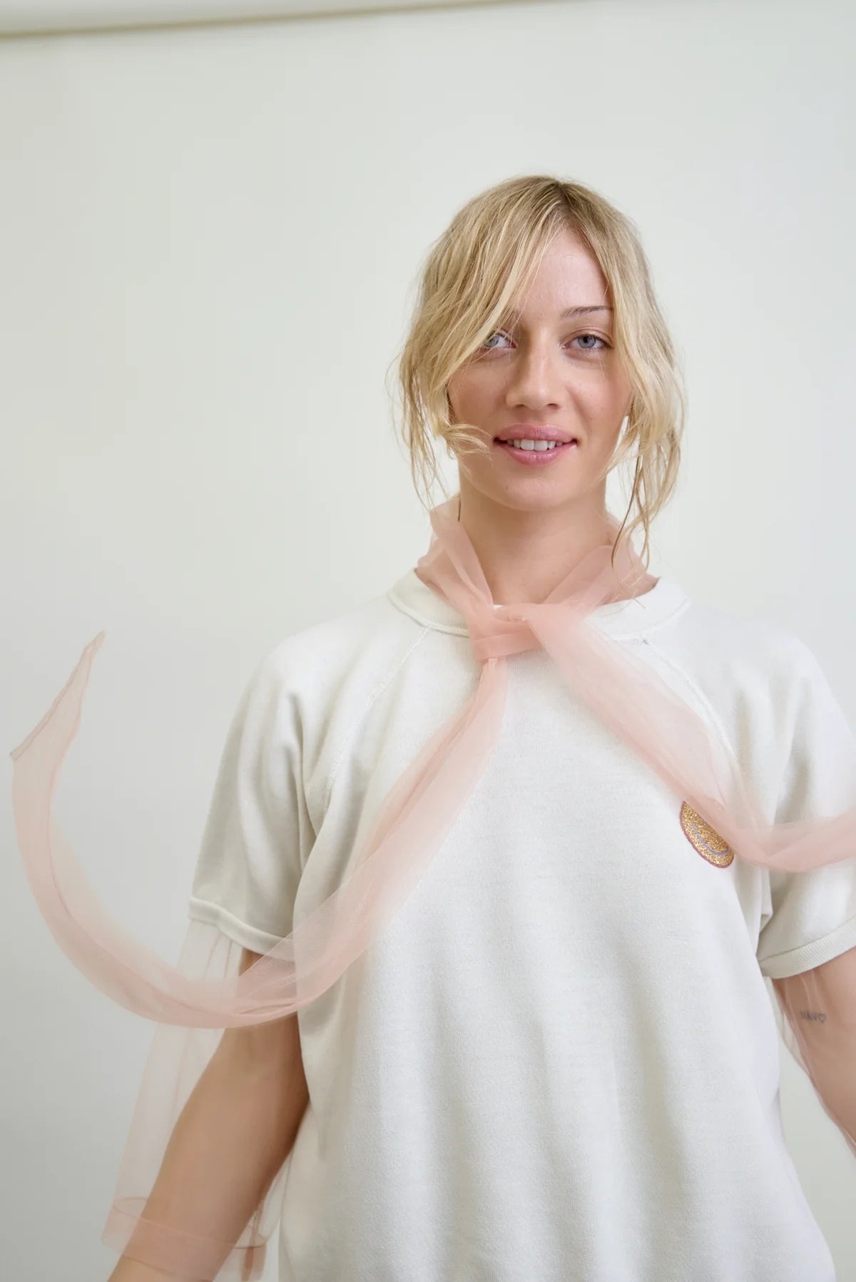 A woman with blonde hair wears an Aquarius Cocktail Dear Polly Bow Blouse against a plain background. A sheer pink scarf is tied around her neck, its ends flowing like mesh tulle. She appears relaxed and smiles gently at the camera.