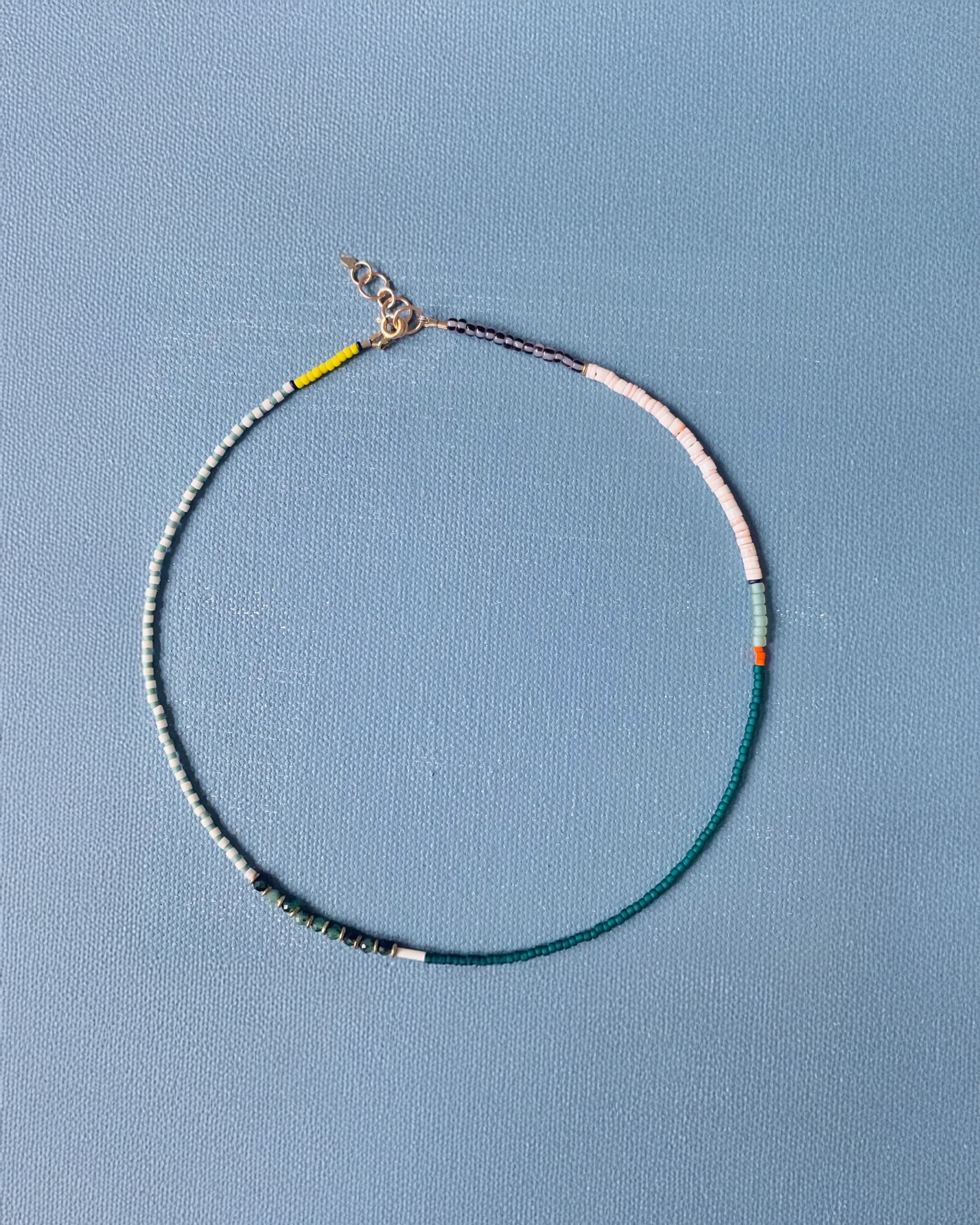 The AR Emerald Necklace by Faire features patterned green, white, yellow, pink, orange, and black beads with a 14k gold-filled clasp, shown on a textured light blue background.