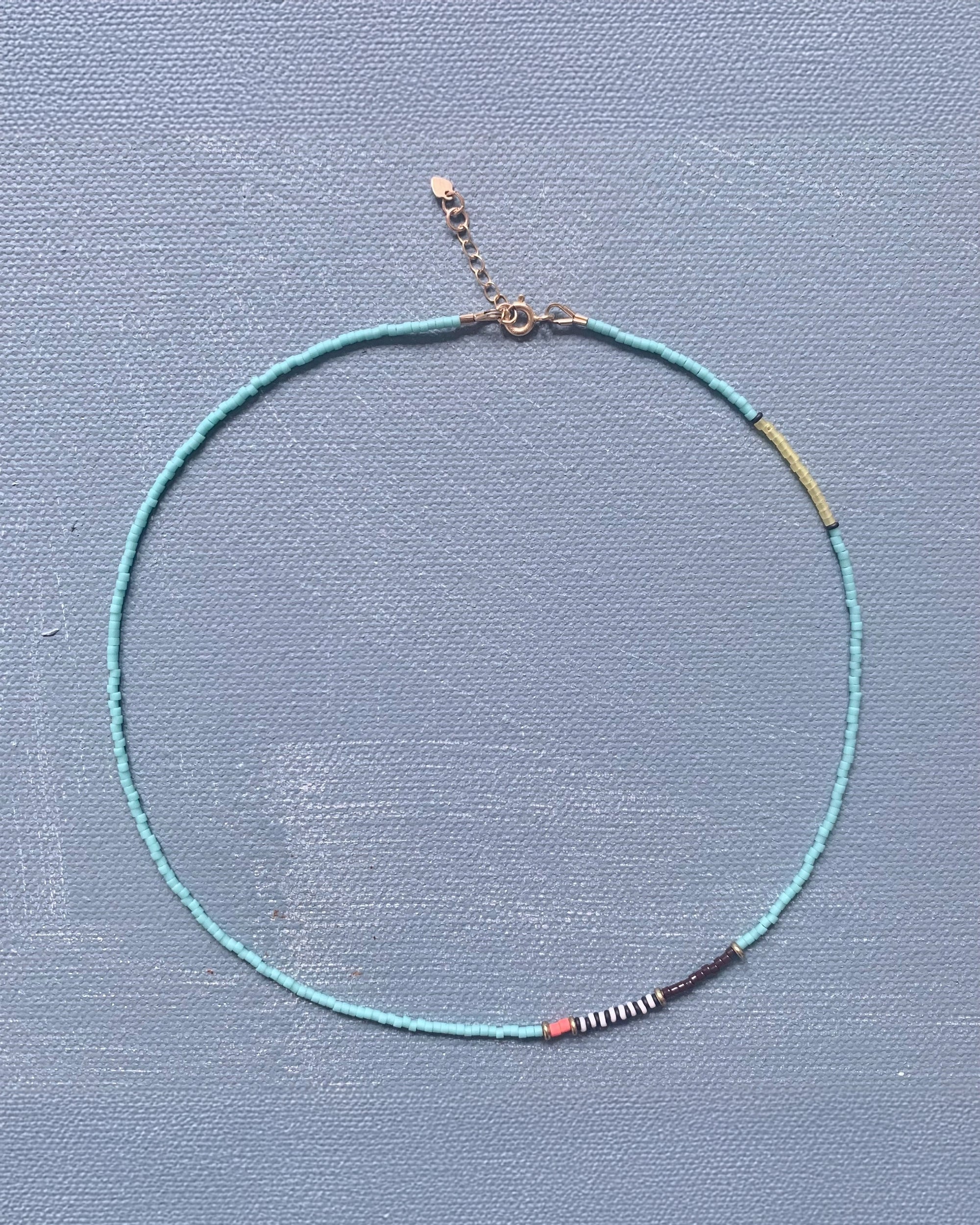 The AR Line Necklace In Turquoise by Faire is a lightweight piece made with small turquoise Japanese glass beads and features black, white, red, and yellow accents, all displayed on a textured light blue background.