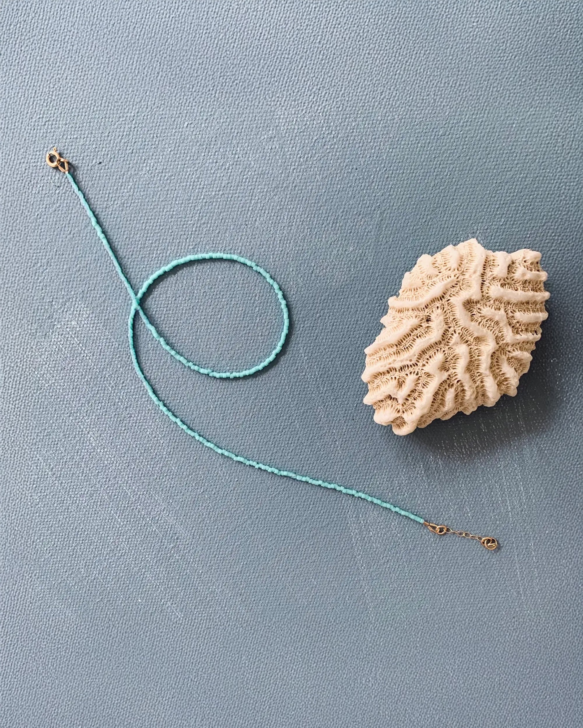 A delicate teal braided bracelet with gold clasps spirals beside beige coral on a light blue surface, pairing beautifully with Faire’s Single Note Beaded Necklace Turquoise.