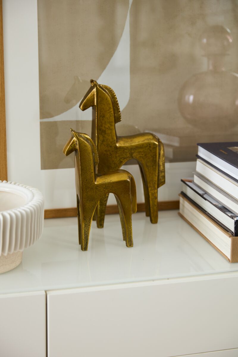 Two Accent Decor Arabian Figurine Small sculptures in antique gold are displayed on a white surface beside a stack of books, a ceramic decorative bowl, and a framed abstract artwork in the background.