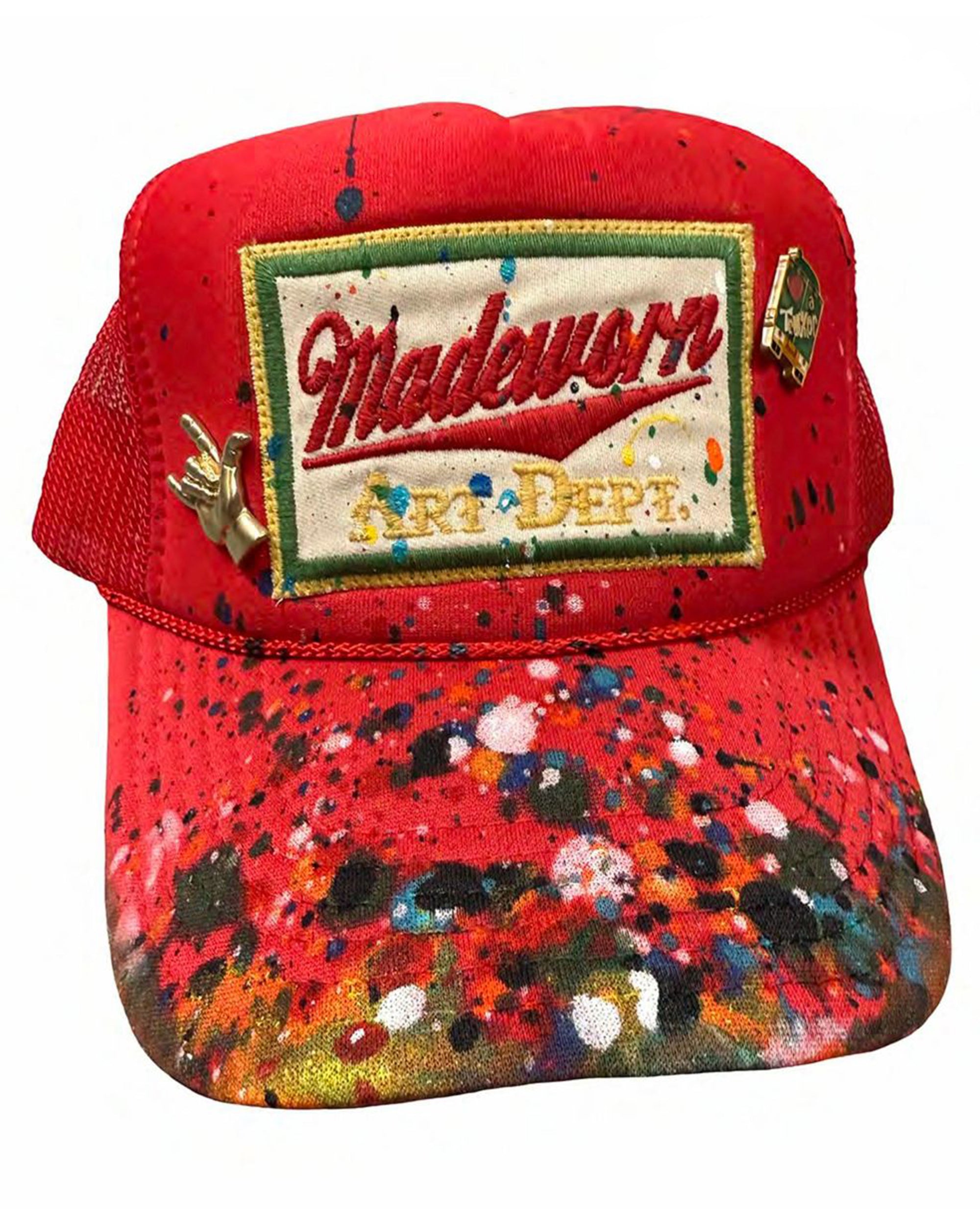The MadeWorn Hat by Made Worn is a red trucker hat with a mesh back, colorful paint splatters, a custom "Madeworn ART DEPT." front patch, and limited edition pins including a gold hand and small circular pin on the front.