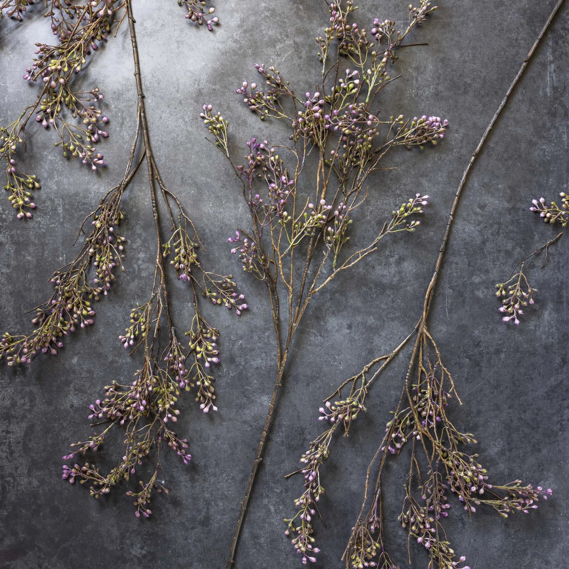 The Berry Branch Lilac by Abigail Ahern features delicate brown branches with small lilac and green artificial berries, arranged on a dark textured surface for a natural, artistic display.