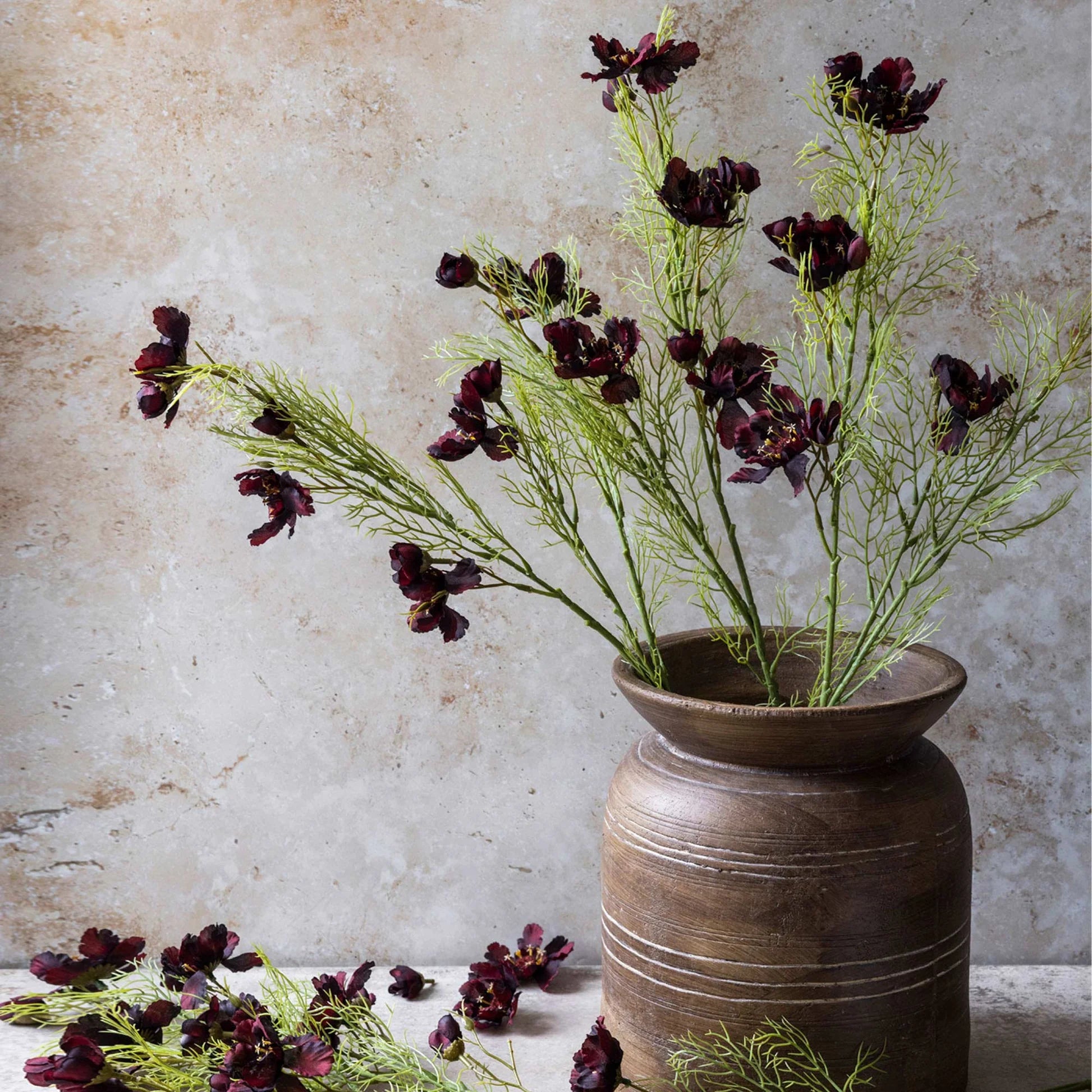 Abigail Ahern’s Cosmos/Burgandy features long green artificial stems with dark burgundy blooms in a rustic brown ceramic vase. Hand-painted fabric flowers are arranged on a light surface, set against a textured beige wall.