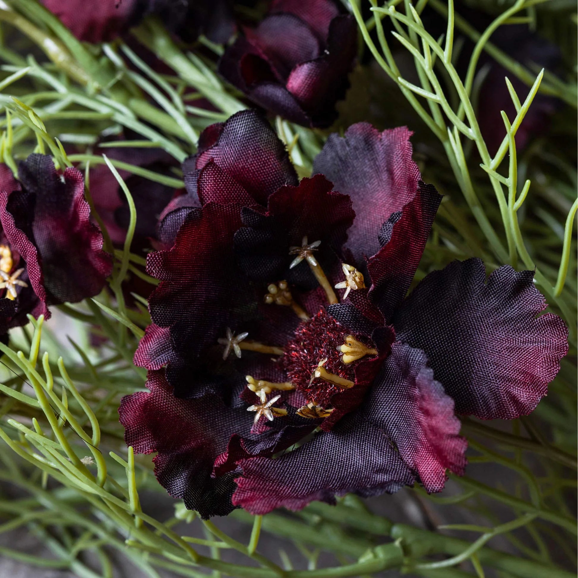 A close-up of Abigail Ahern's Cosmos /Burgandy artificial flower stem features textured dark burgundy petals, yellow stamens, and thin green grass-like foliage.