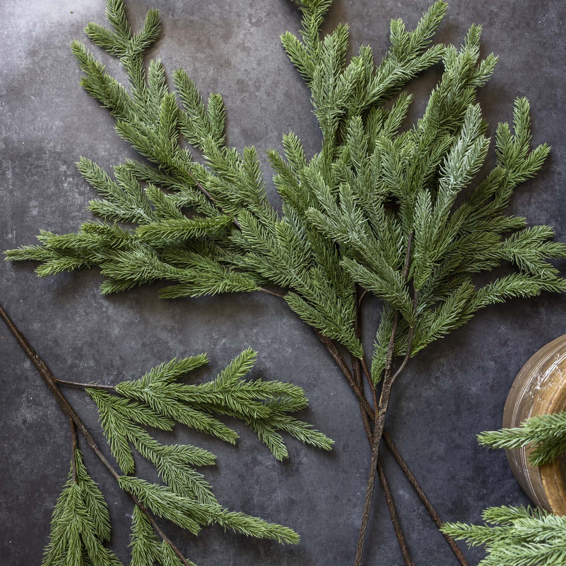 Sprigs of green, needle-like Abigail Ahern Foxtail Pine branches are displayed on a dark gray stone surface, with a portion of a round wooden object visible in the lower right corner.