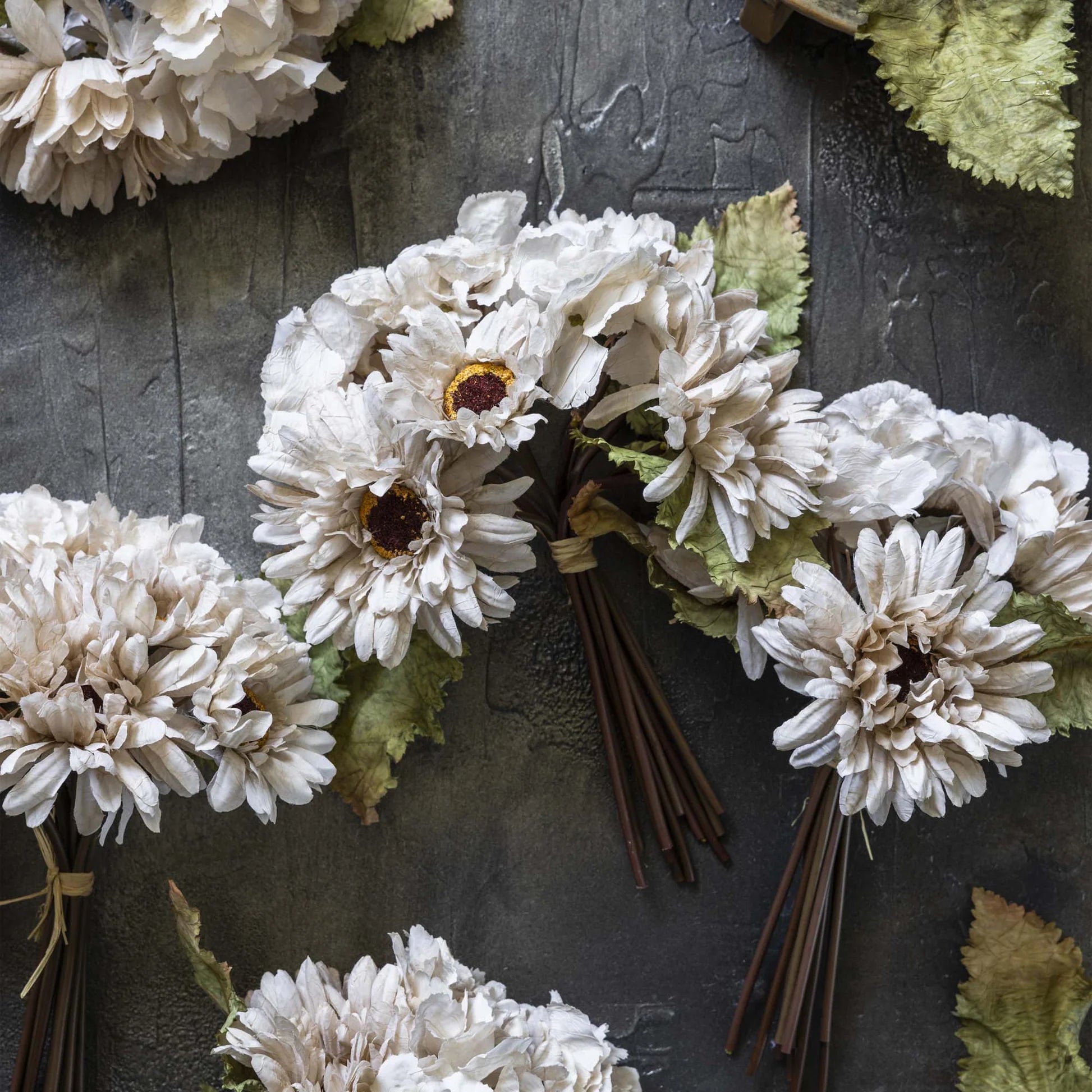 Several small hand-painted fabric bouquets from Abigail Ahern's AA-N4-001 Garden Bouquet, featuring dark centers and green leaves, are tied with twine and arranged on a textured dark gray surface.