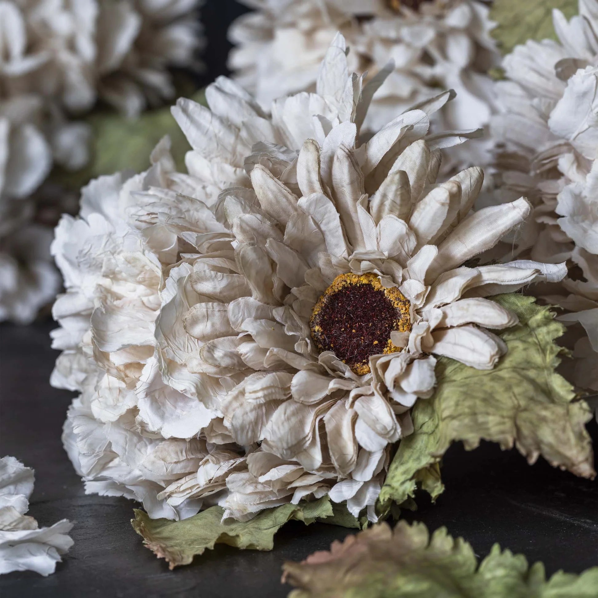 Close-up of AA-N4-001 Garden Bouquet by Abigail Ahern: a white, ruffled hand-painted fabric flower with a dark brown and yellow center, surrounded by green textured leaves. More faux flower stems appear softly blurred in the background.