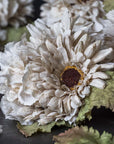 Close-up of AA-N4-001 Garden Bouquet by Abigail Ahern: a white, ruffled hand-painted fabric flower with a dark brown and yellow center, surrounded by green textured leaves. More faux flower stems appear softly blurred in the background.