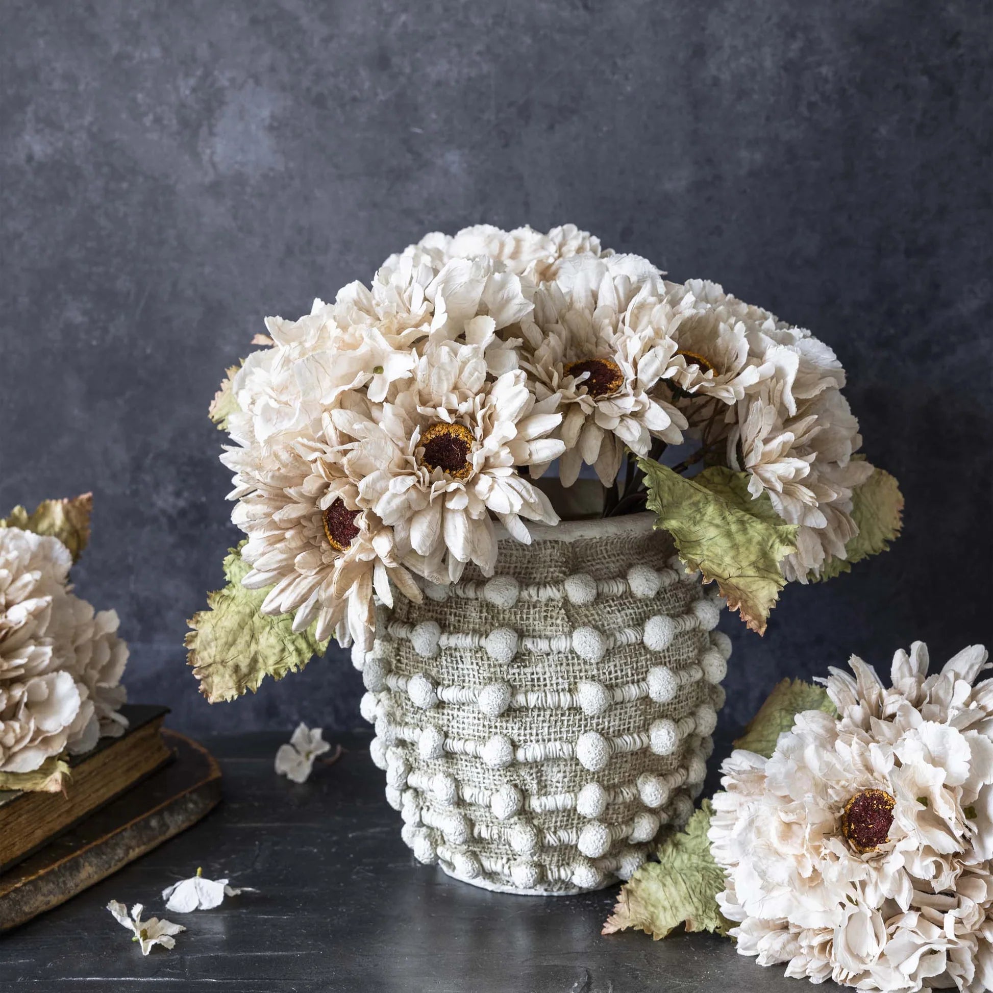 The Abigail Ahern AA-N4-001 Garden Bouquet, a textured white vase with cream hand-painted fabric flowers and greenish leaves, is displayed on a dark surface with extra faux stems and an old book against a dark gray background.