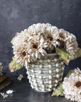 The Abigail Ahern AA-N4-001 Garden Bouquet, a textured white vase with cream hand-painted fabric flowers and greenish leaves, is displayed on a dark surface with extra faux stems and an old book against a dark gray background.