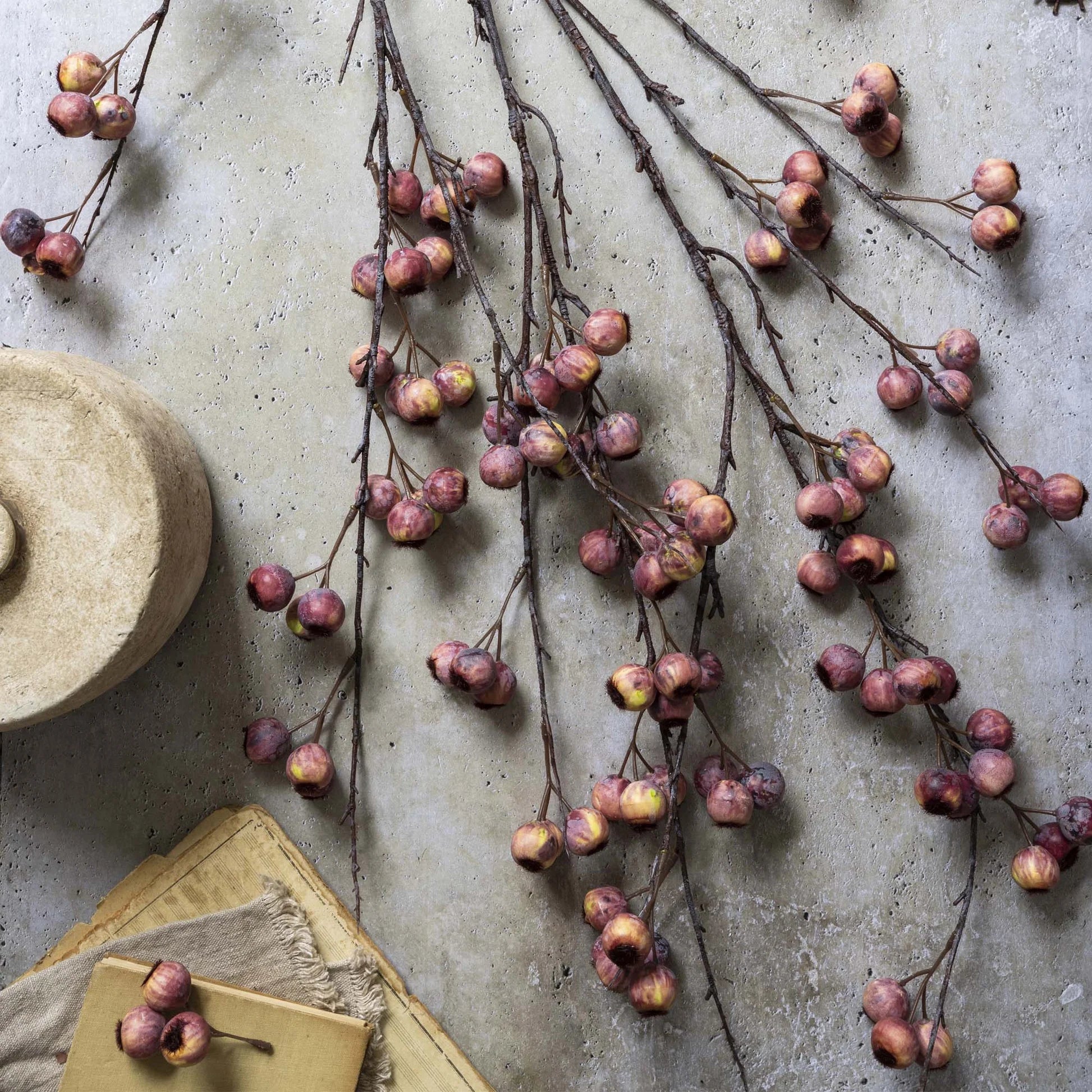 Abigail Ahern's Hawthorne Berry branches, adorned with clusters of small purple and yellow berries, are displayed on a textured stone surface beside a beige container and neatly stacked cloth and paper in the corner.